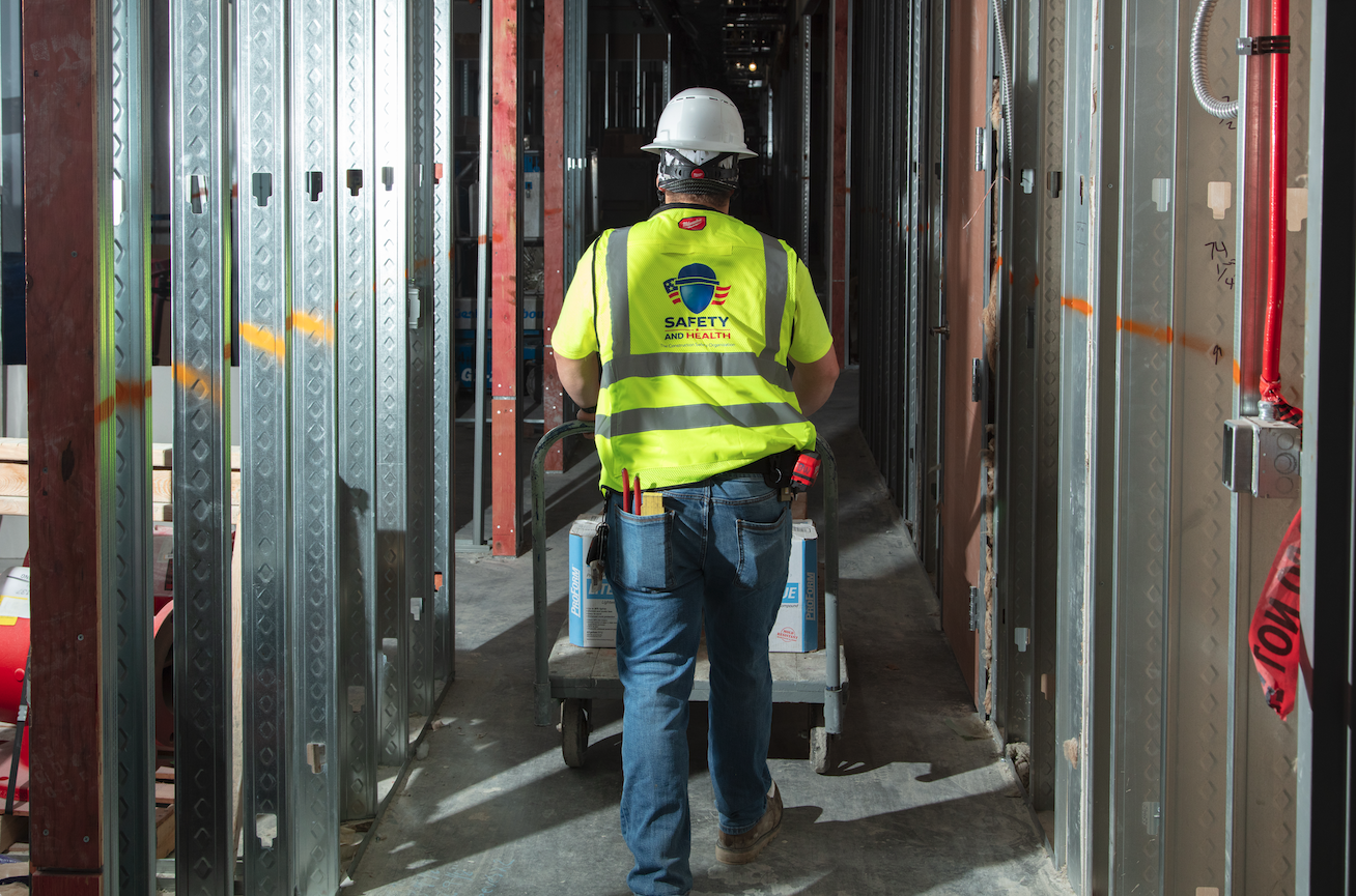 A construction worker wearing a yellow safety vest and white hard hat walks with a cart inside a building under construction, surrounded by metal framing for walls.