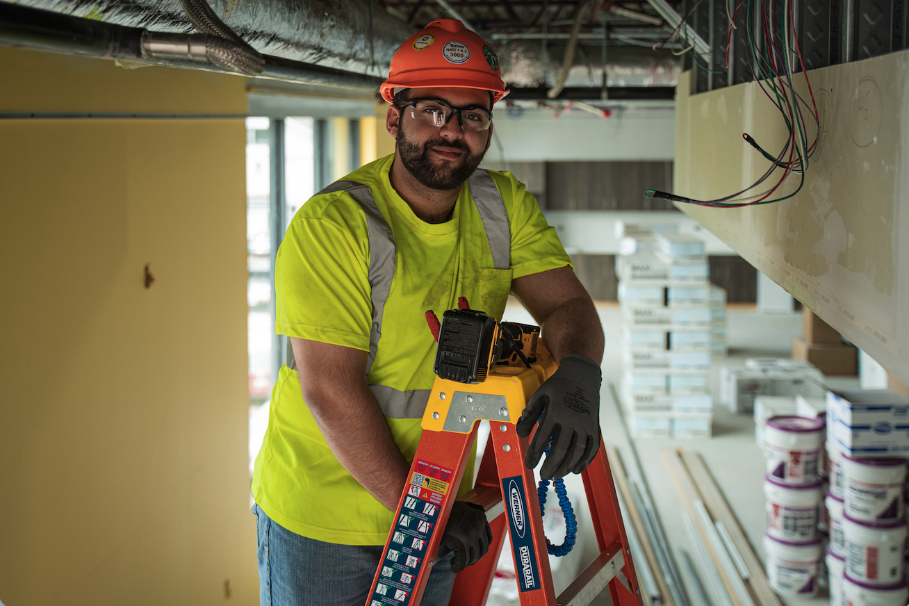 A construction worker with a beard, glasses, and a yellow safety shirt, wearing an orange helmet, standing indoors on a ladder, working with electrical wiring.