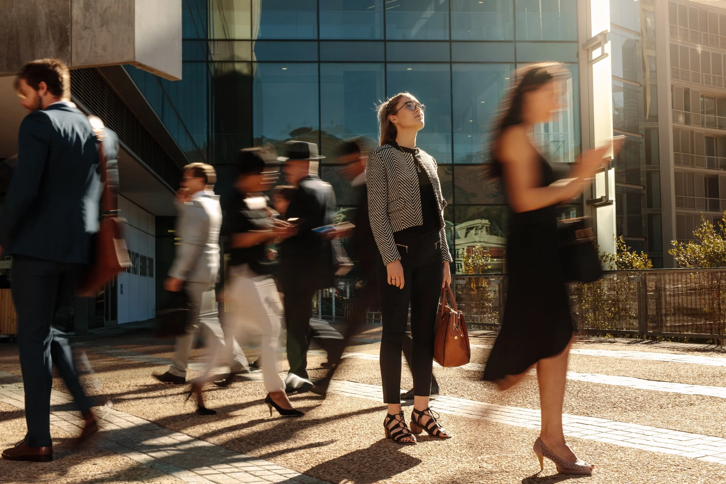 A woman with glasses and a black and white patterned blazer stands still in a busy city street, surrounded by pedestrians walking past her, some blurred due to movement, with modern glass buildings in the background and sunlight casting warm tones.