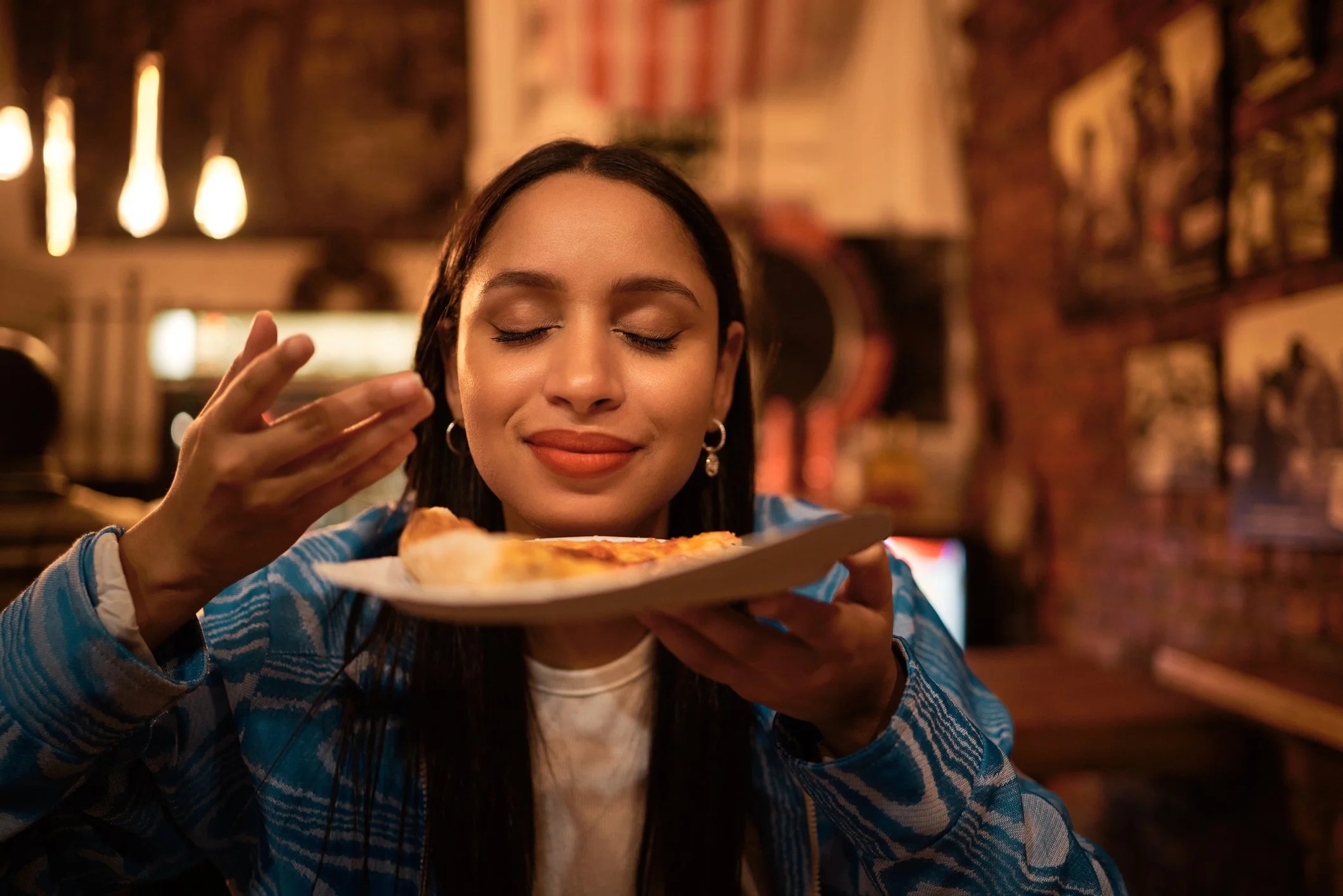 A woman with closed eyes and a smile, wearing hoop earrings and a blue patterned jacket, is holding a slice of pizza on a plate near her face in a cozy, warmly lit restaurant.