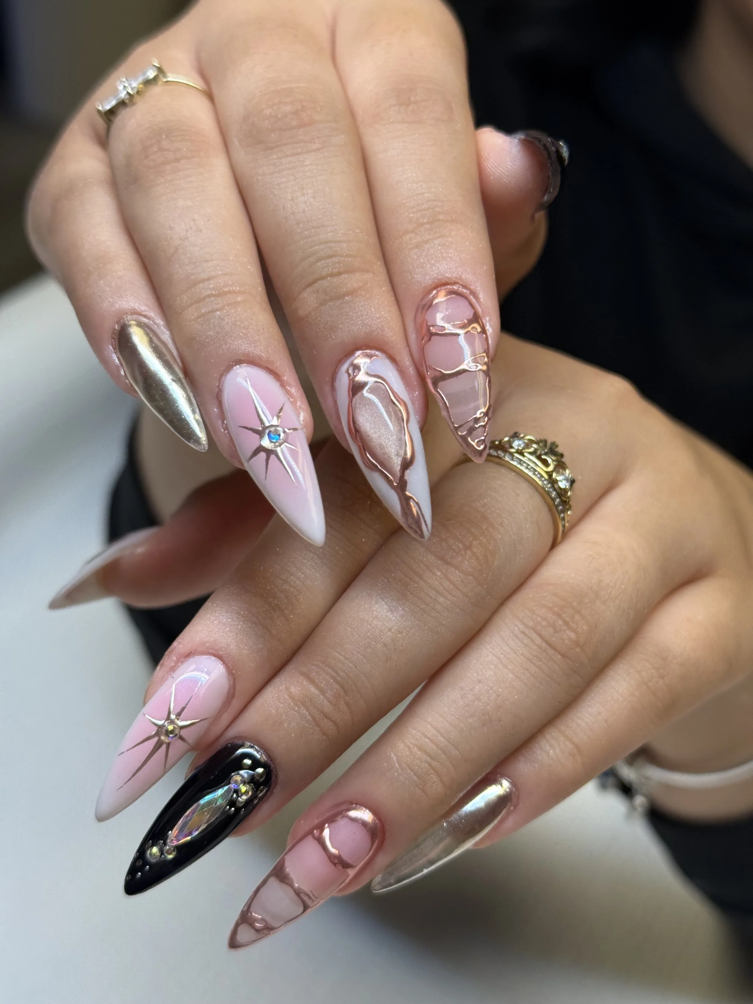 Close-up of a person's hand with long, almond-shaped nails featuring intricate metallic, pink, black, and iridescent nail art designs, some with rhinestone embellishments. The person is wearing rings on their fingers.