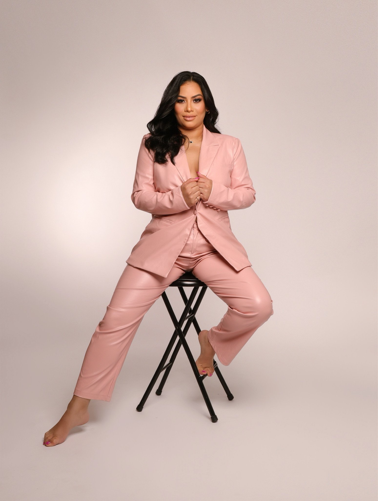 A woman in a light pink satin pantsuit sitting on a black stool against a plain light background. She has long dark hair, tan skin, and is barefoot with pink nail polish.