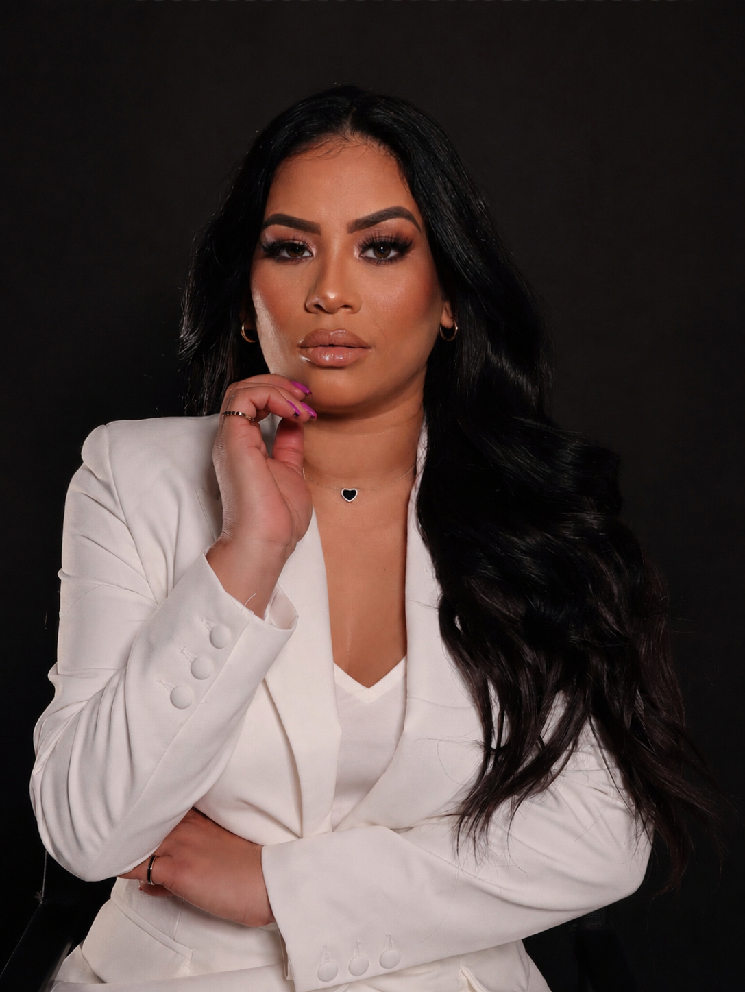 A woman with long black hair, wearing a white blazer and a white top, poses with her hand near her chin against a dark background.