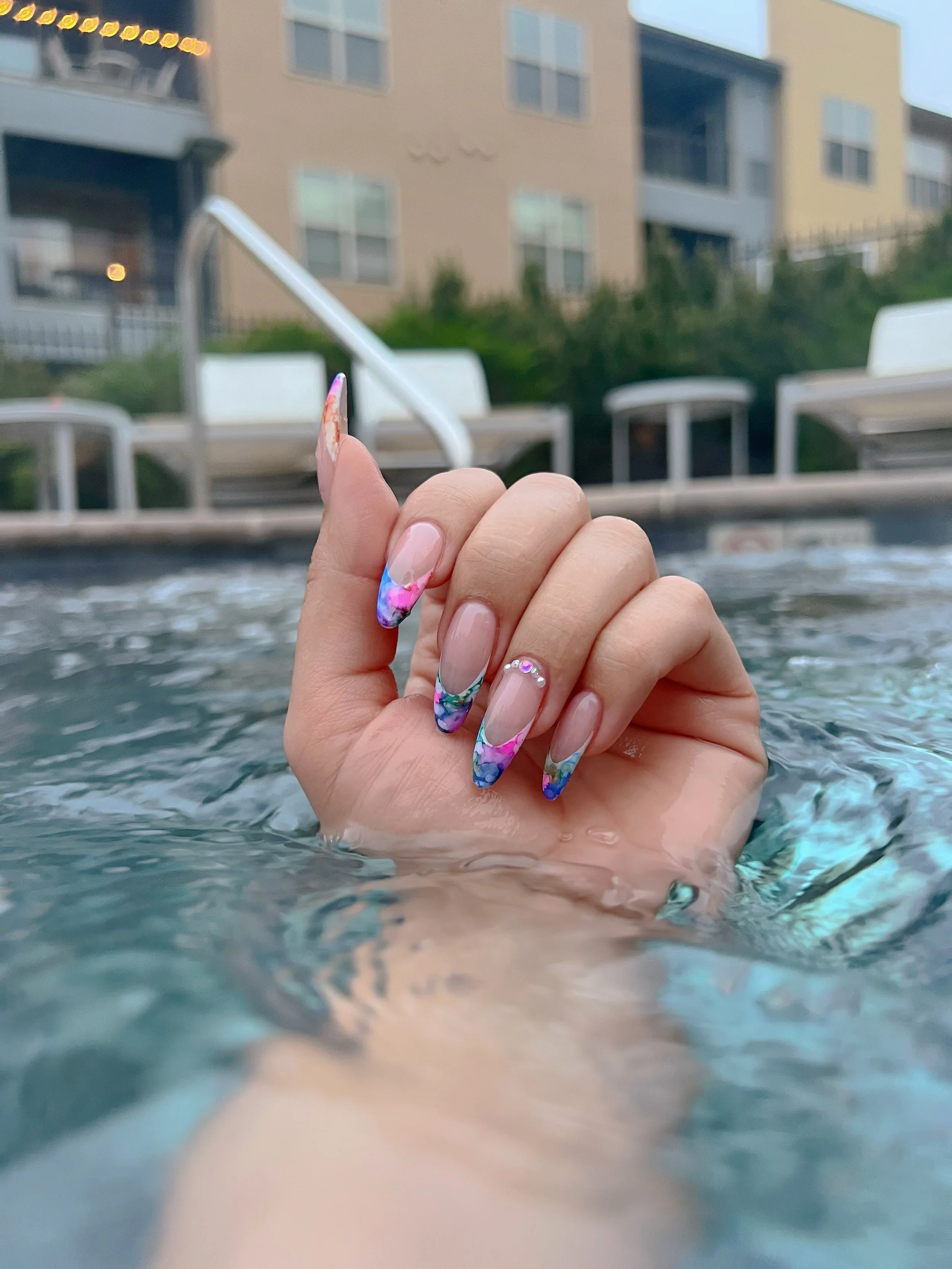 Person with long manicured nails in colorful marble design, holding onto a handrail in a pool with apartment buildings in the background.