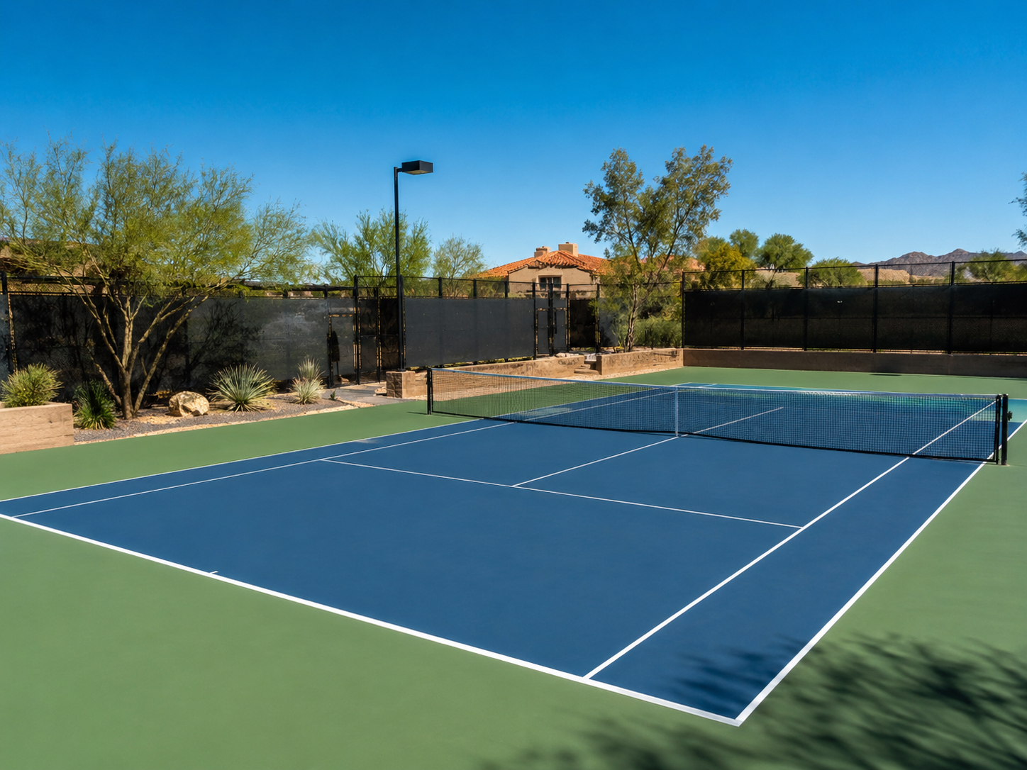An outdoor tennis court with blue playing surface and white lines, surrounded by desert landscaping and a black fence, under a clear blue sky.