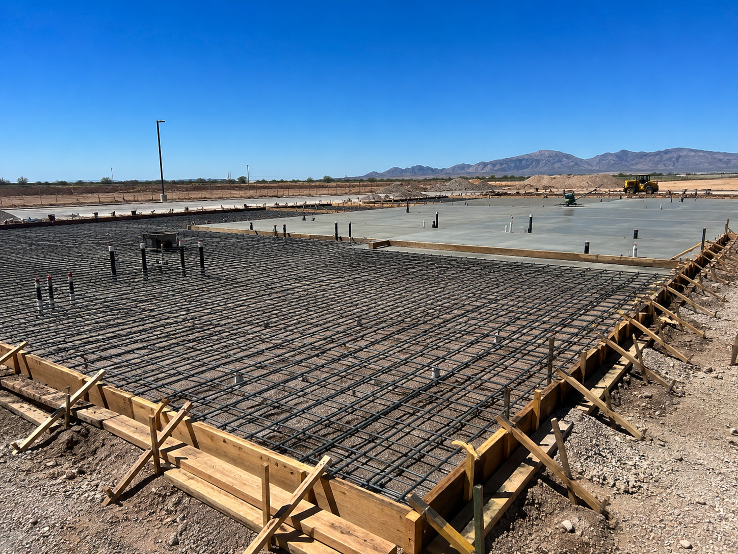 Construction site with concrete foundation and steel reinforcement bars laid out, surrounded by wooden formwork, in a desert landscape with mountains in the background.