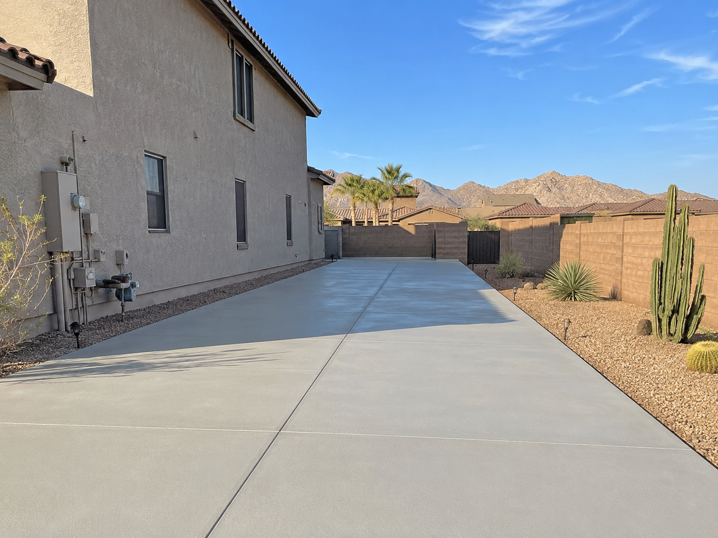 A concrete backyard patio with desert plants, cacti, and a brown privacy fence, with mountains in the background under a blue sky.