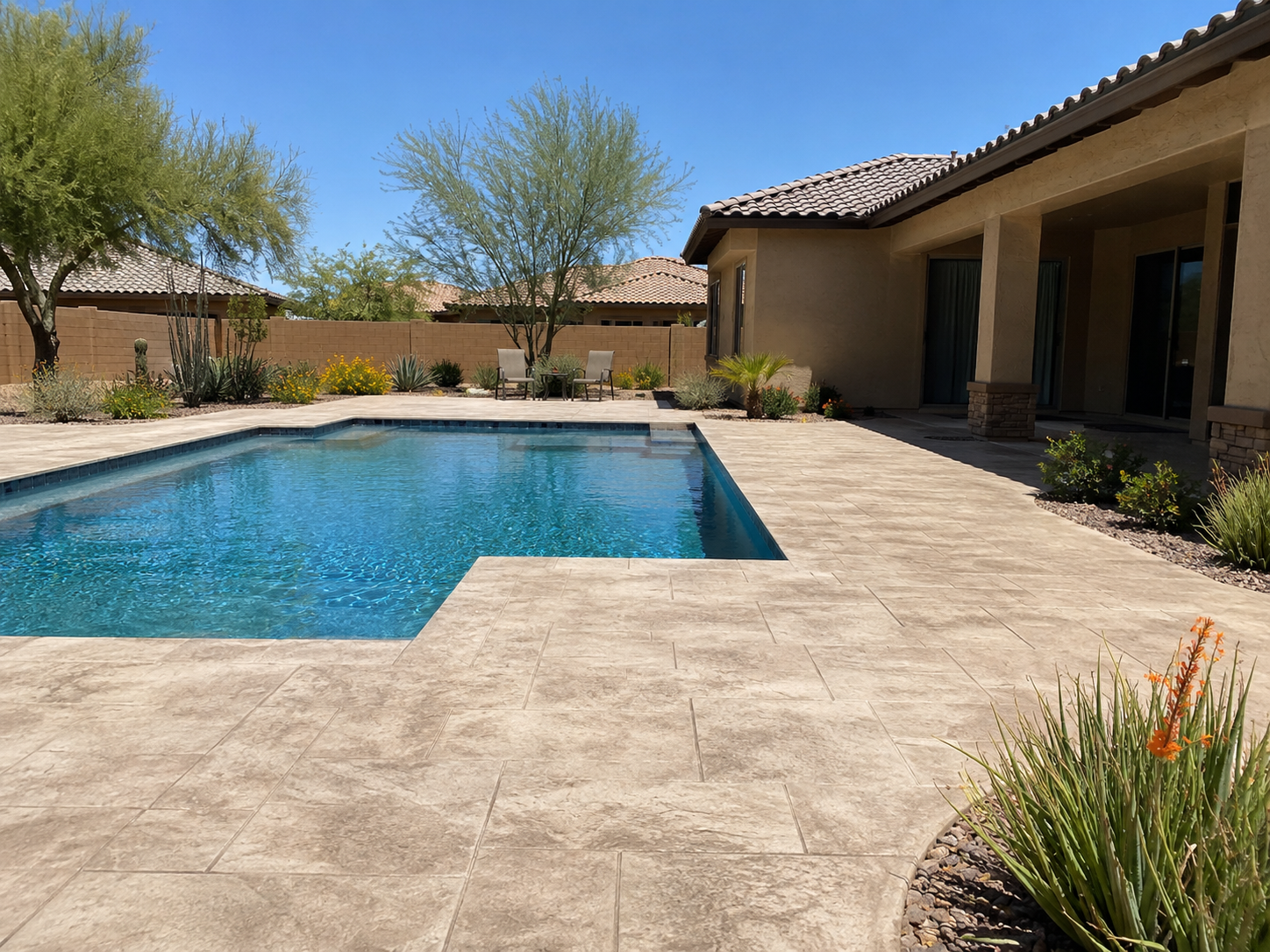 A backyard with a blue swimming pool surrounded by a beige stone patio, with desert plants and shrubs, a house with beige walls and a tiled roof, and chairs and trees in the background under a clear blue sky.