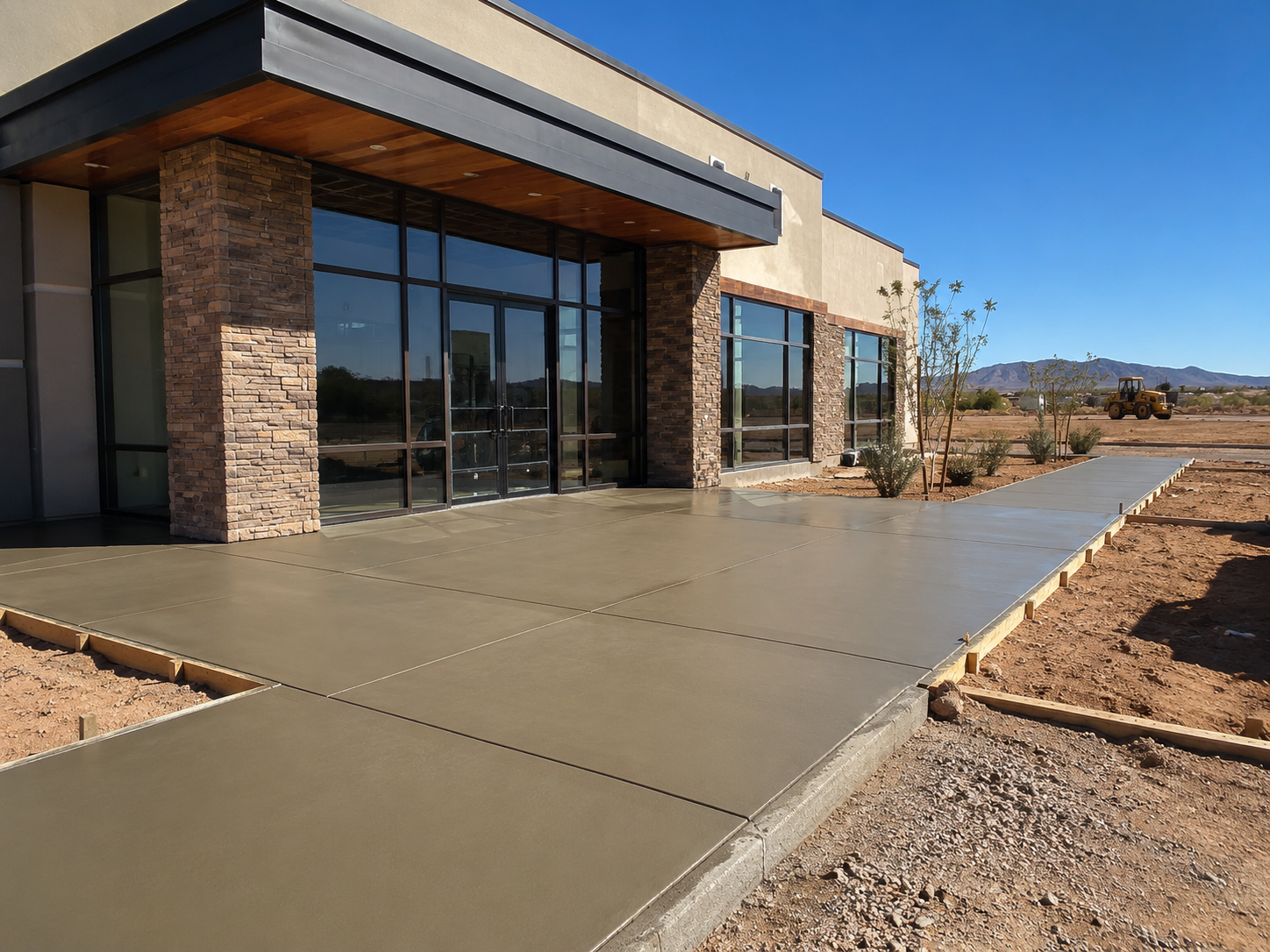 Newly poured concrete sidewalk in front of a modern commercial building with glass windows and brick accents, in a desert landscape.