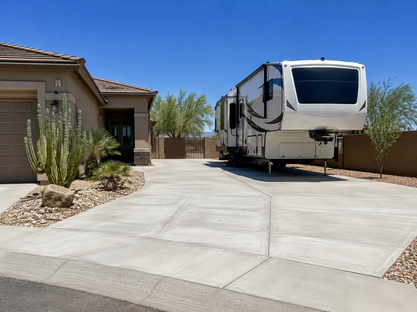 A large white fifth-wheel RV parked on a concrete driveway next to a desert-themed front yard with cacti and small trees in a suburban neighborhood under a clear blue sky.