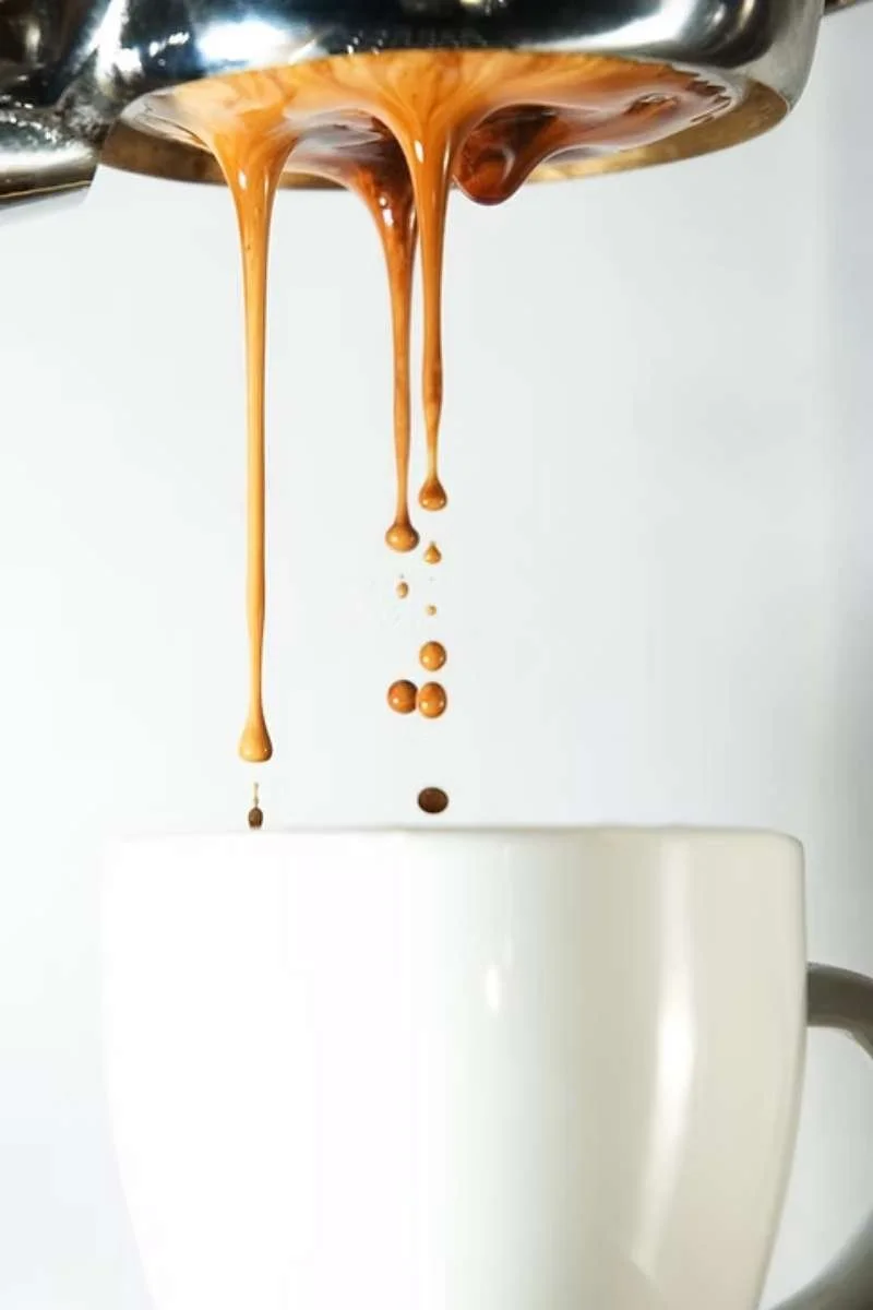 Hot espresso coffee flowing from a portafilter into a white mug, with a plain light background.