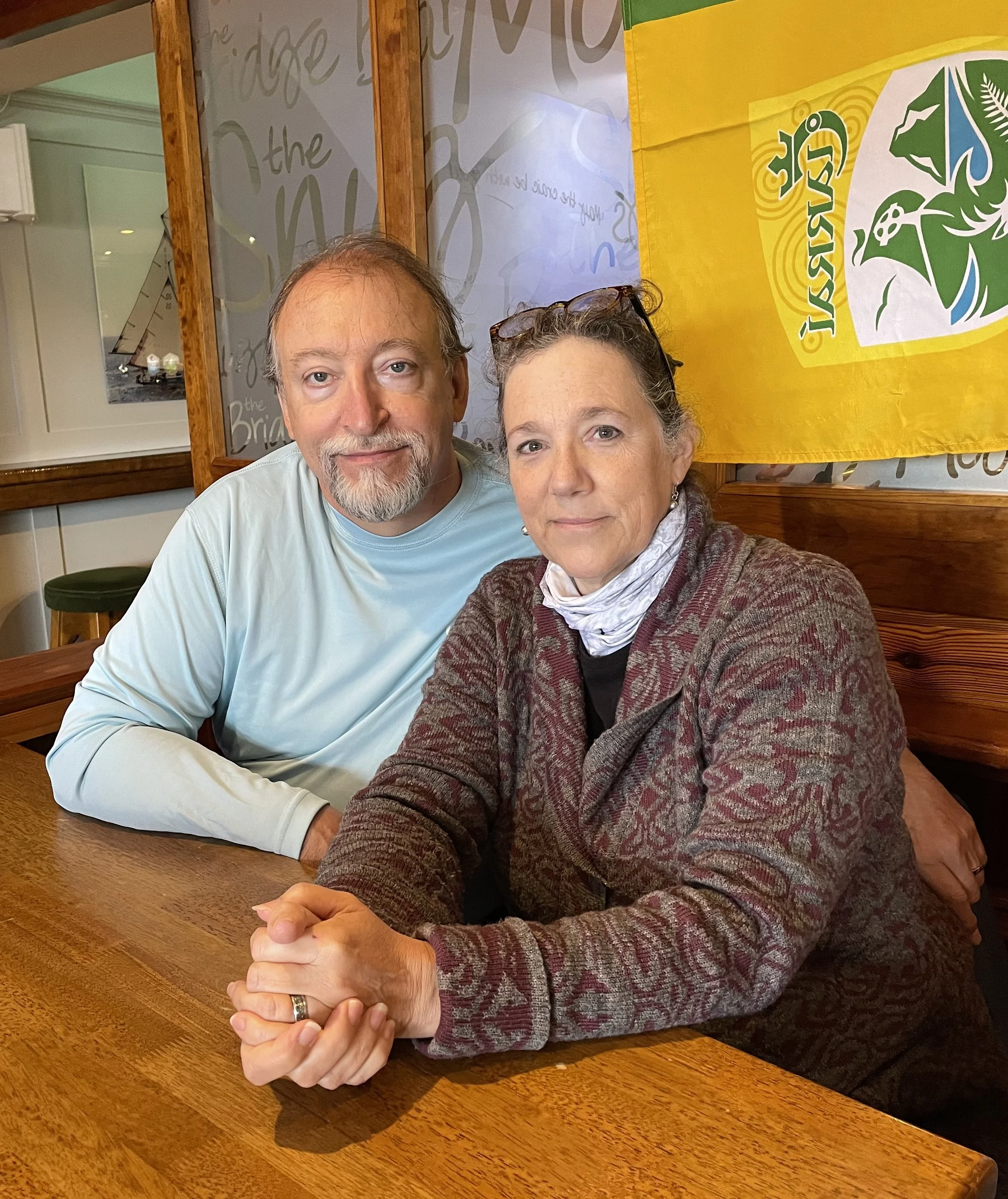 A middle-aged man and woman sitting at a wooden table, holding hands and looking at the camera, with a yellow and green Jamaica flag hanging in the background.