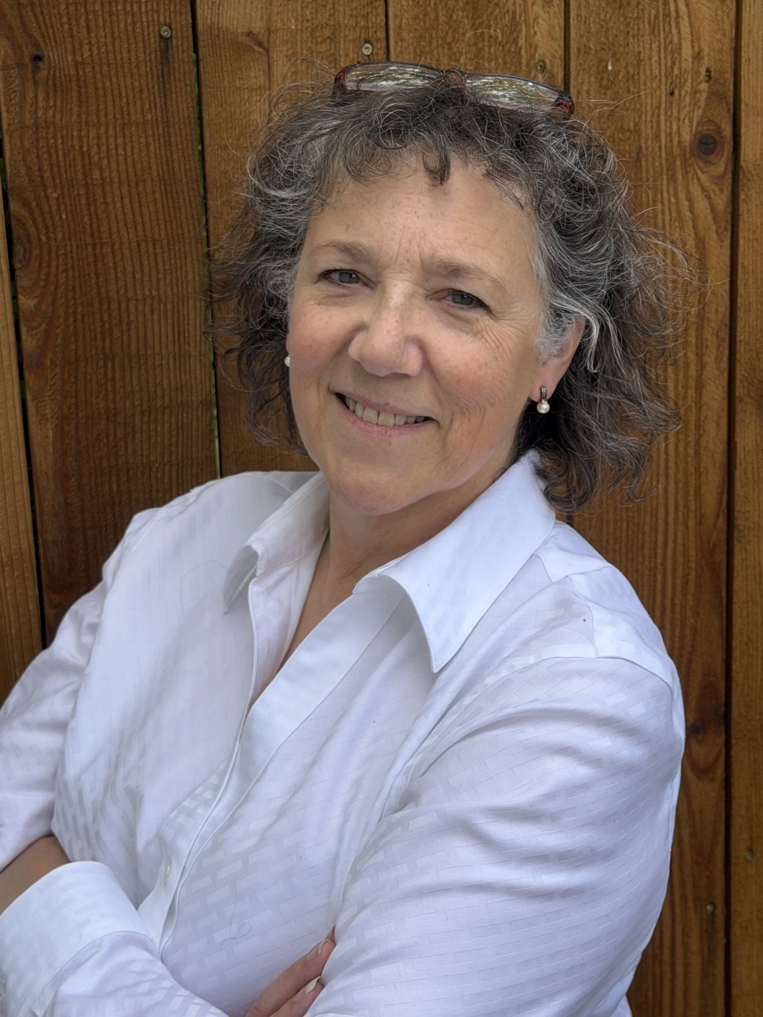 A middle-aged woman with curly, gray hair and glasses resting on her head is smiling and crossing her arms, standing in front of a wooden fence.
