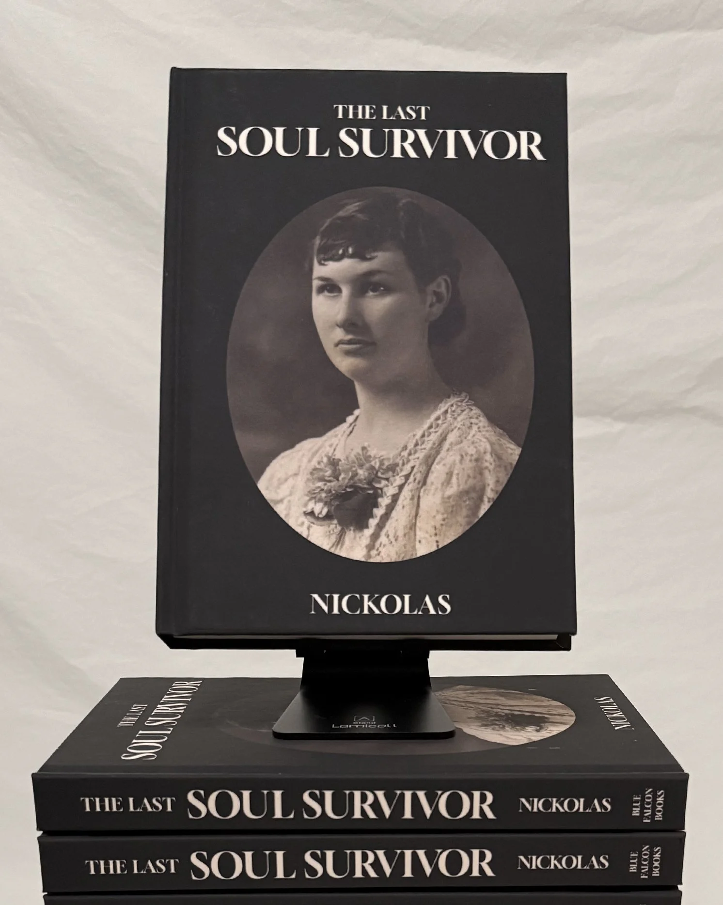 A black hardcover book titled "The Last Soul Survivor" by Nickolas features a photograph of a woman with short dark hair, wearing a lace dress and a flower brooch, on the cover. The book is standing vertically on top of several copies of the same book, which are stacked horizontally on a flat surface, with some additional promotional material around it.