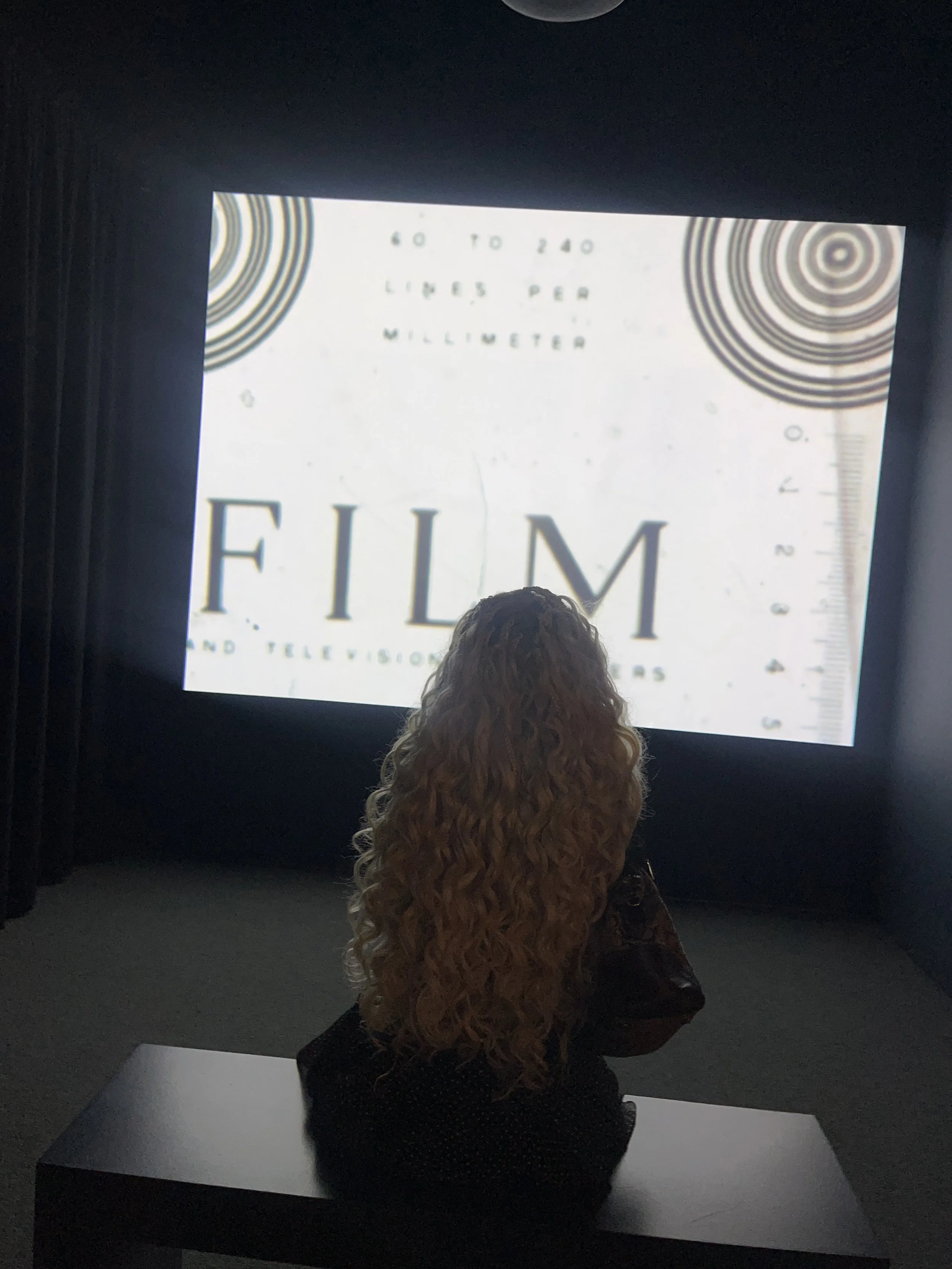 Person with curly hair sitting at a table in front of an illuminated screen displaying text and designs, with dark surroundings.