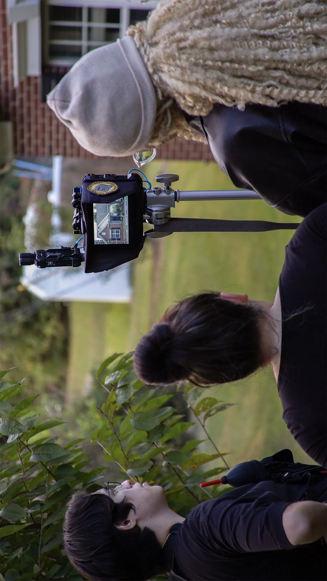 Two women outdoors, one woman filming with a camera on a tripod, the other woman looking at plants, in a backyard or garden with grass and trees.