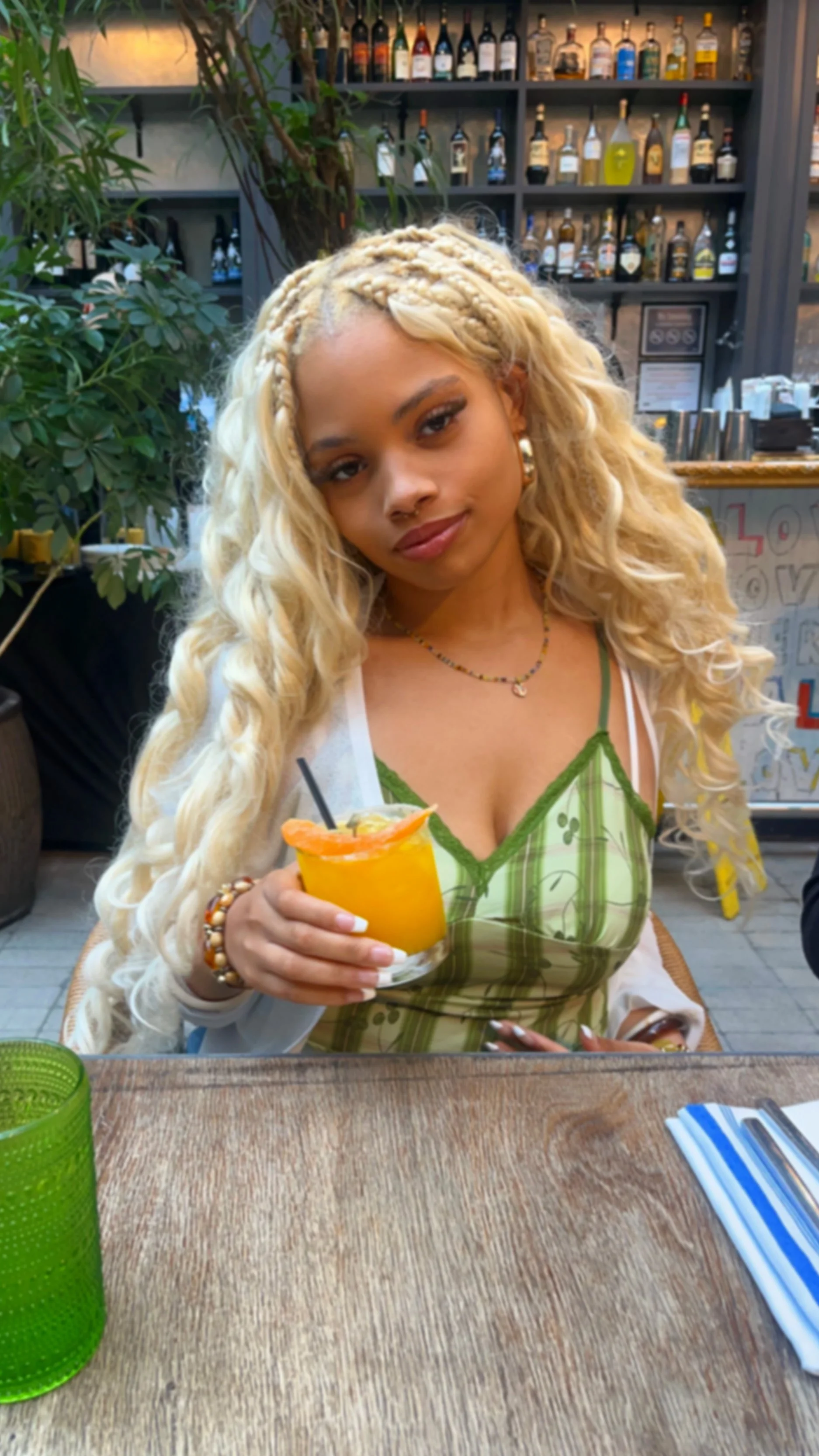 A woman with long, curly blonde hair and a braid, holding a yellow cocktail with an orange slice and a straw, sitting at a wooden table in a restaurant or bar with bottles of alcohol on shelves behind her.