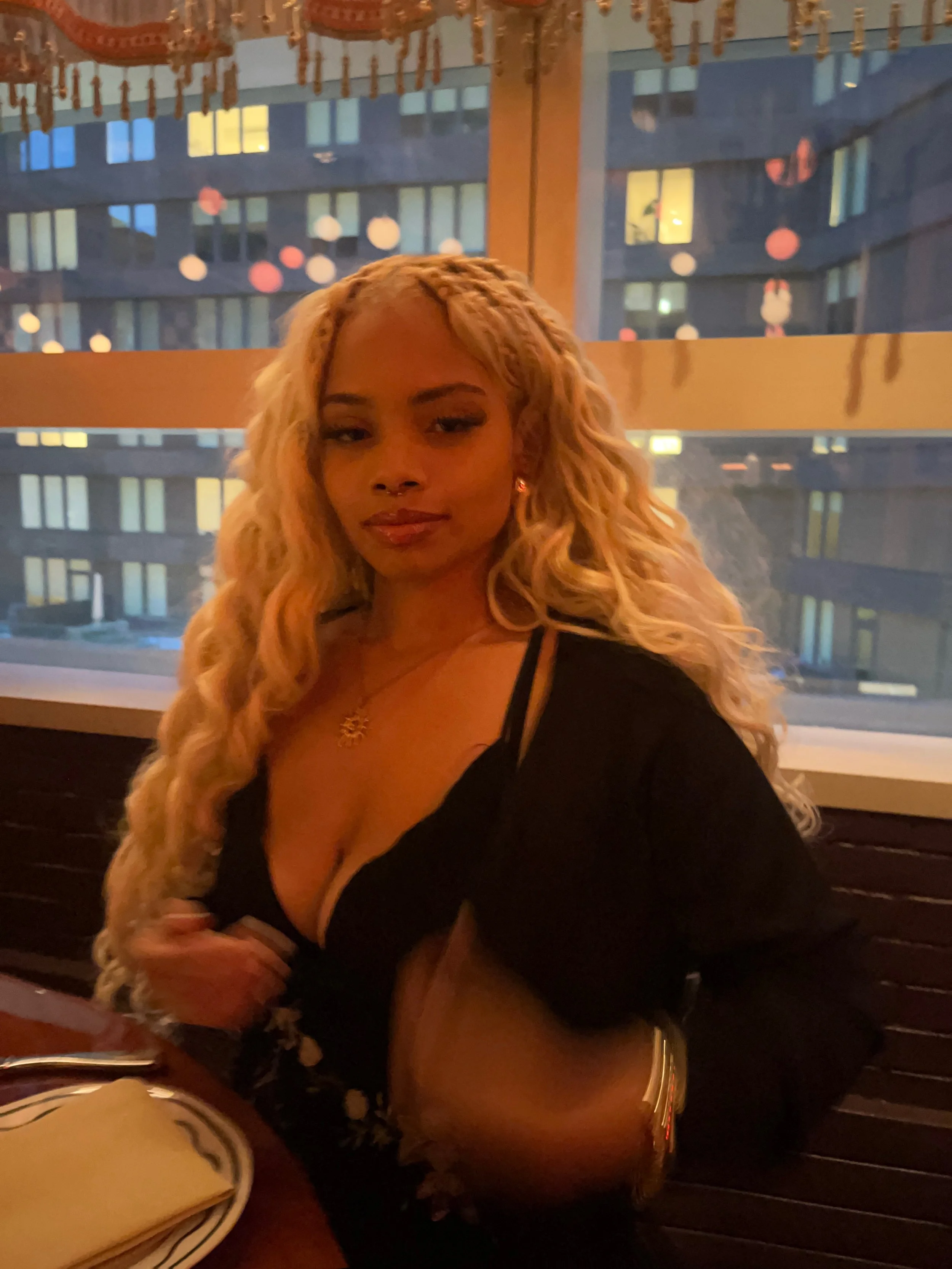 A woman with long, curly blonde hair sitting at a table in a dimly lit restaurant or bar with decorative lanterns hanging from the ceiling and a large window in the background.