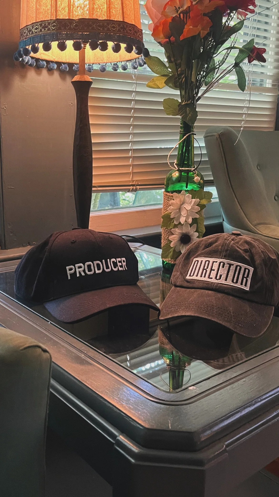 A glass coffee table with two hats labeled "PRODUCER" and "DIRECTOR" on top. Behind the table, a vase with artificial white flowers, a lamp with a decorative shade, and window blinds are visible.