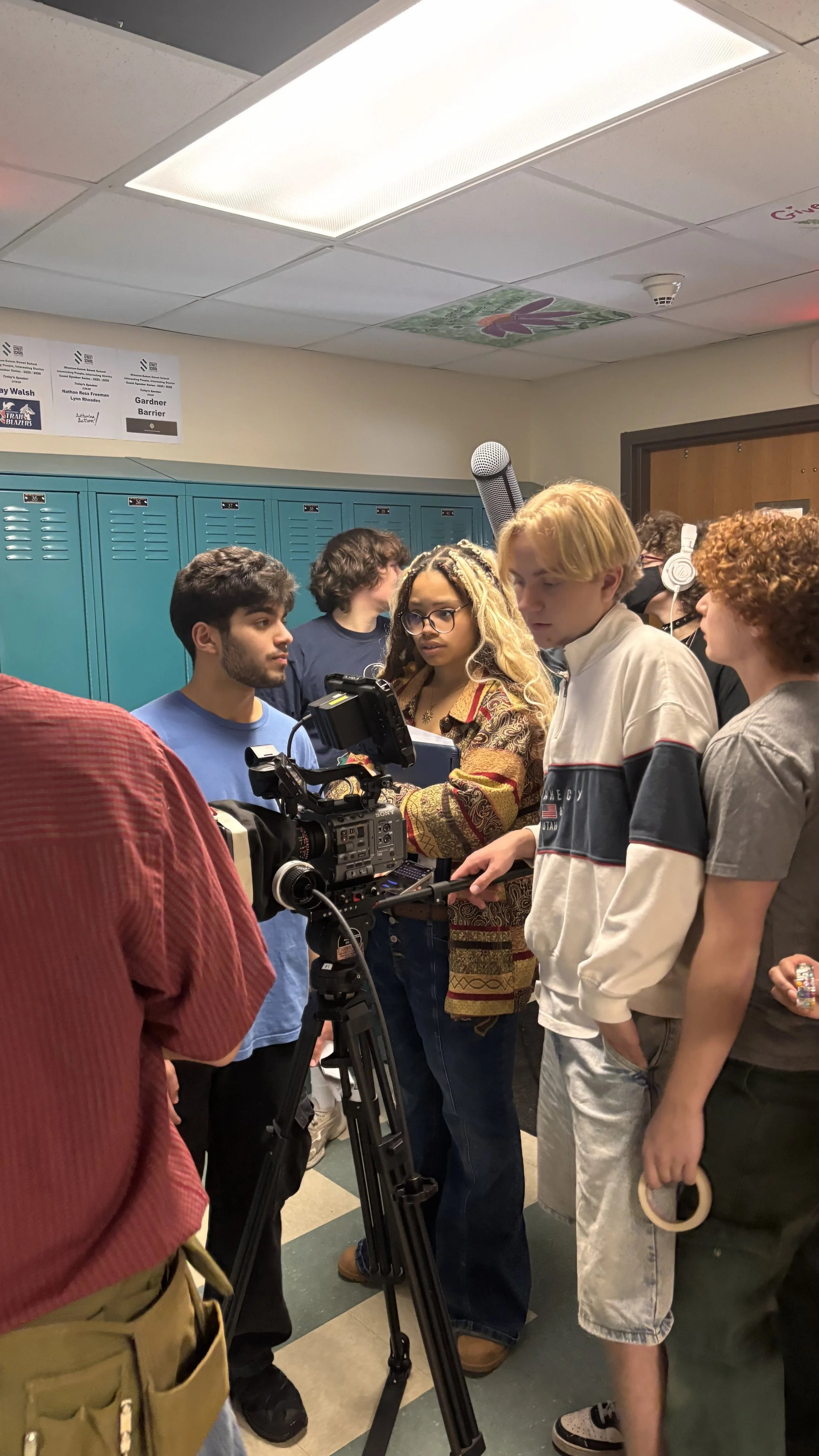 Group of students and a young woman filming with professional camera equipment in a school hallway with teal lockers. Several students are standing around, some wearing headphones.