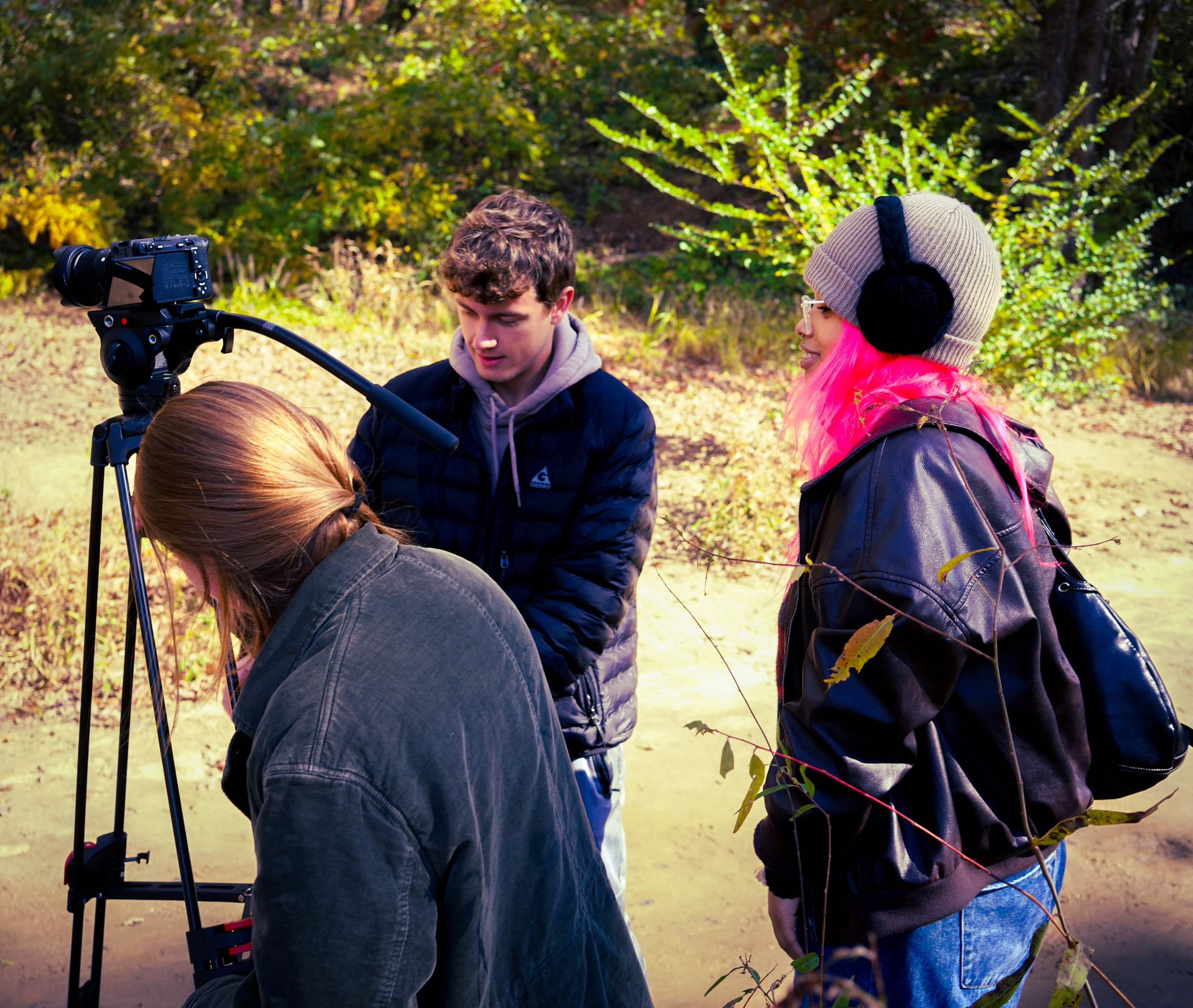 A group of people, including a woman with bright pink hair and a gray knit hat, are outdoors in a park with autumn foliage. They are gathered around a person operating a camera on a tripod, possibly filming or taking a photo.