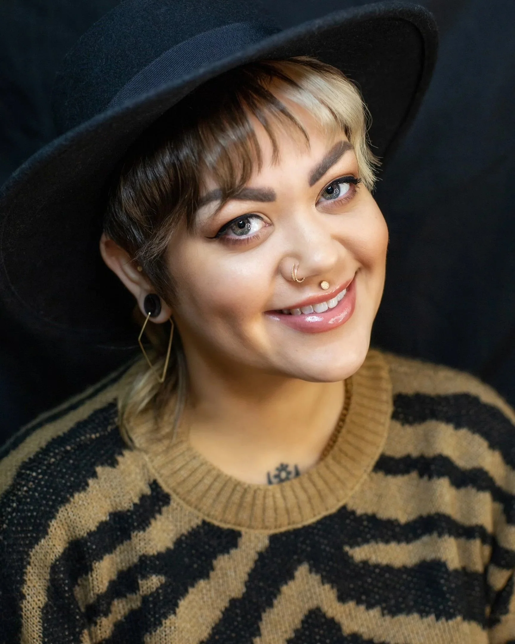 Close-up of a person with short dark and blonde hair, wearing a black wide-brimmed hat, gold nose and ear jewelry, and a brown and black patterned sweater, smiling with blue eyes against a black background.