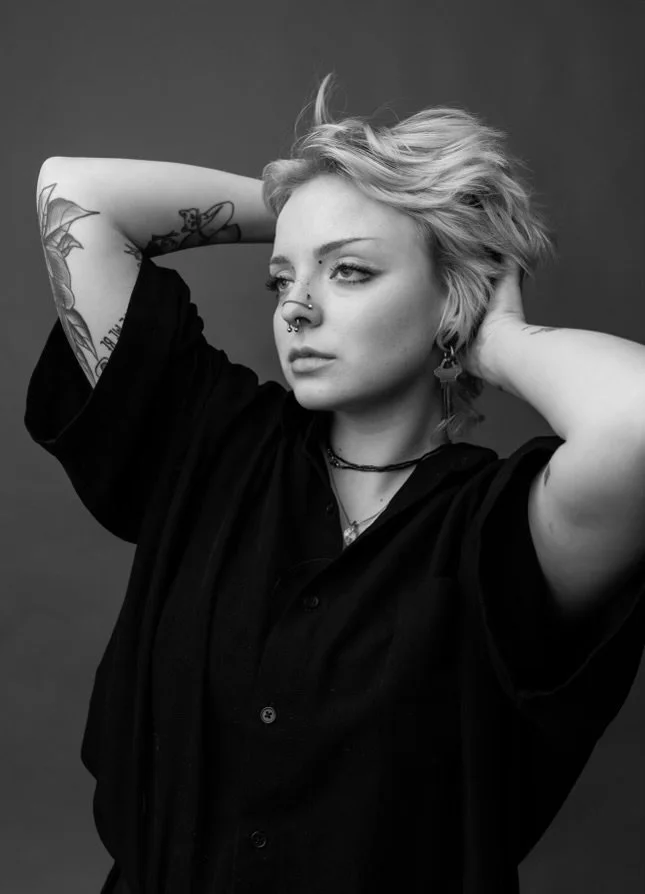 Black and white photo of a young person with short, wavy blonde hair, tattoos on their arms, multiple piercings, wearing a black button-up shirt, posing with hands behind their head against a plain background.