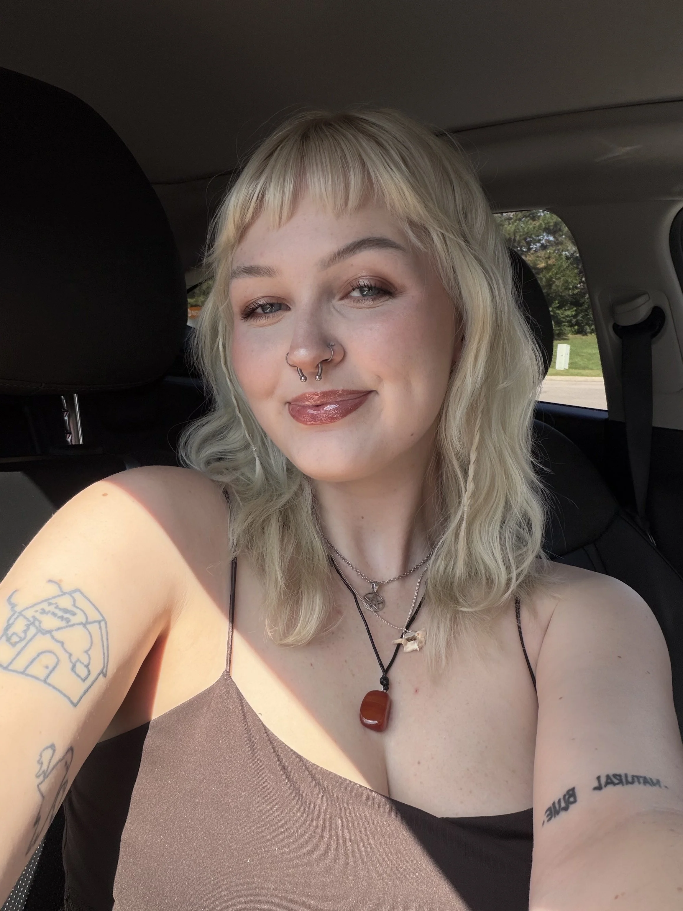 A person with blonde, wavy, shoulder-length hair, blue eyes, multiple piercings including septum, nostril, and earlobes, wearing makeup with winged eyeliner and glossy lips, dressed in layered jewelry and a black hoodie, sitting in a vehicle.