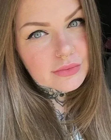 Close-up portrait of a person with long red hair, blue eyes, nose rings, and tattoos visible on their neck.