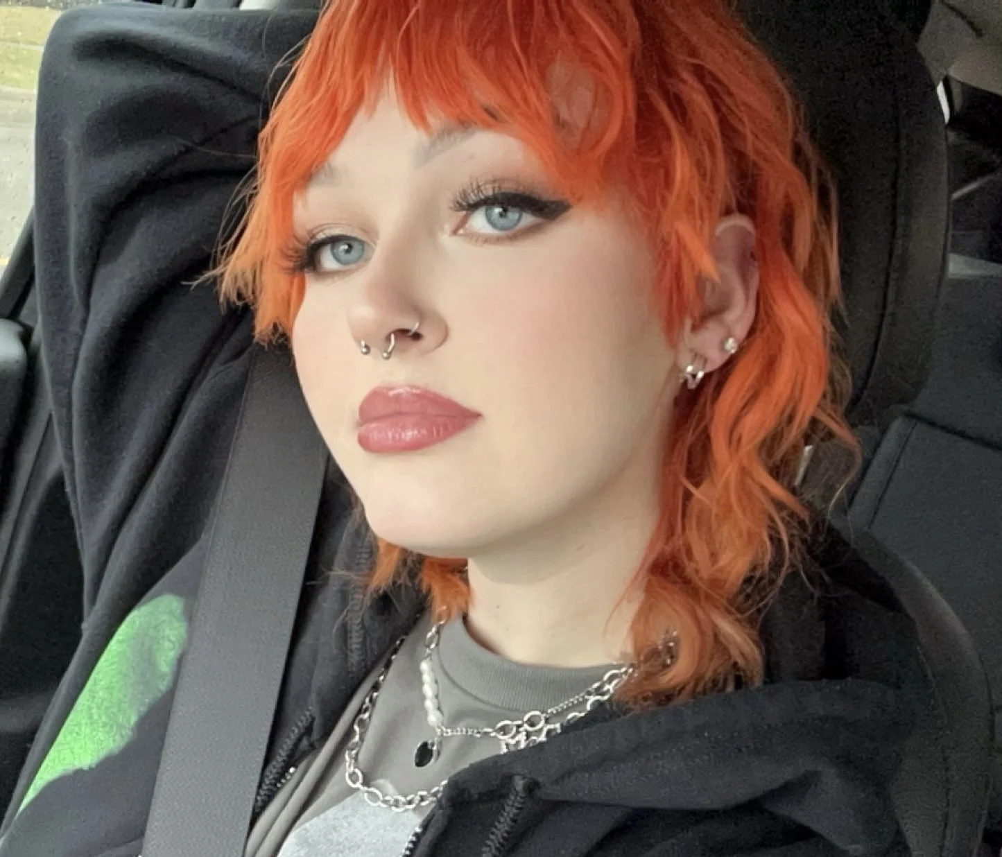 A person with bright orange, wavy, shoulder-length hair, blue eyes, multiple piercings including septum, nostril, and earlobes, wearing makeup with winged eyeliner and glossy lips, dressed in layered jewelry and a black hoodie, sitting in a vehicle.