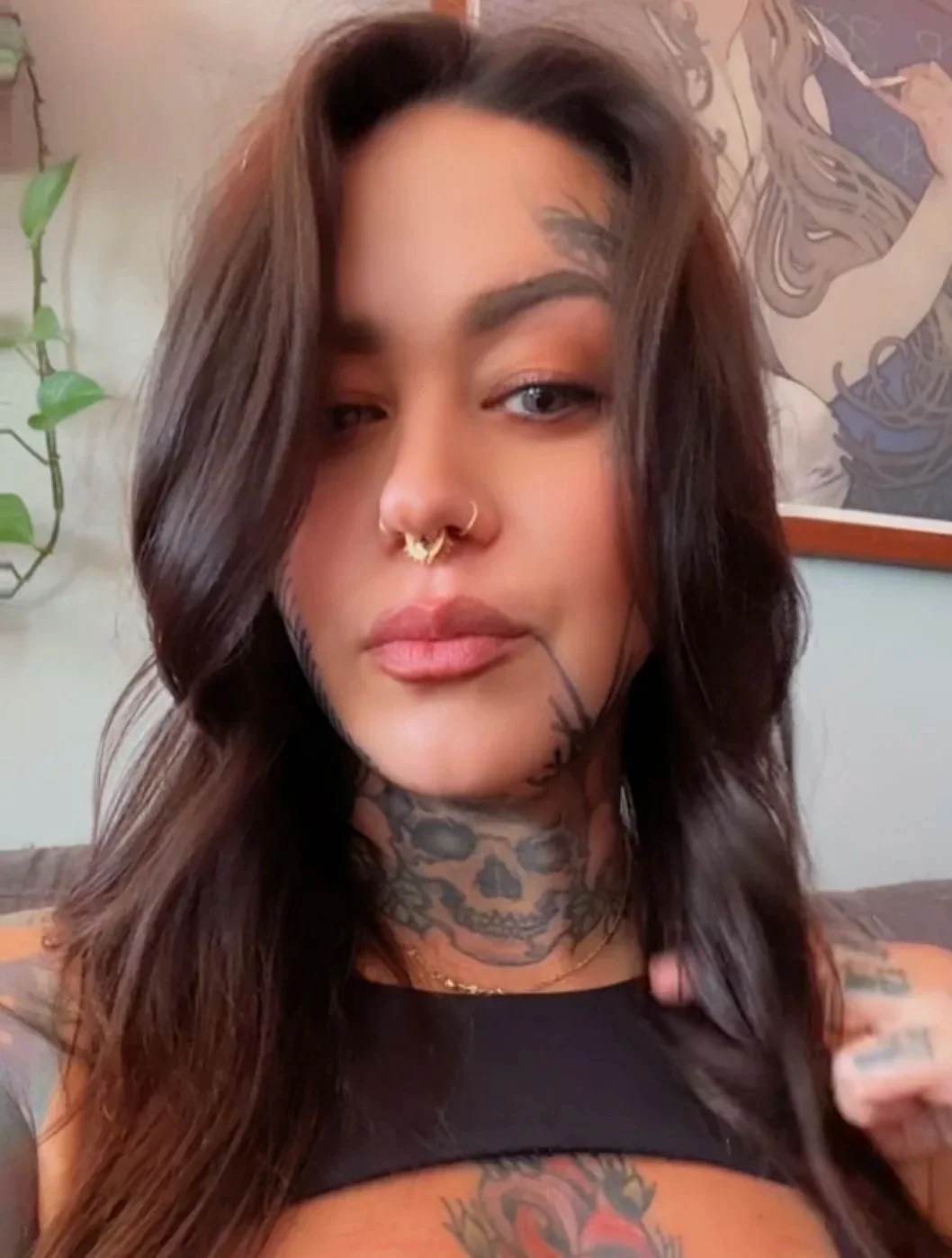 A person with long dark hair, tattoos on their neck and shoulder, wearing a Septum piercing and a black top, with a background featuring a wall with a drawing or mural.