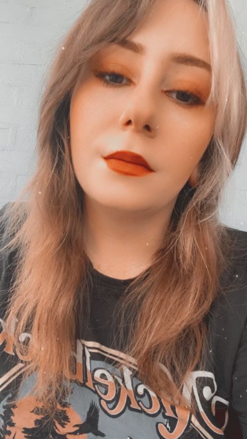 A person with long, wavy light brown hair and fair skin taking a selfie. They have makeup with orange eyeshadow and bold red lipstick, and a silver nose ring. They are wearing a black graphic T-shirt and is standing in front of a white brick wall.