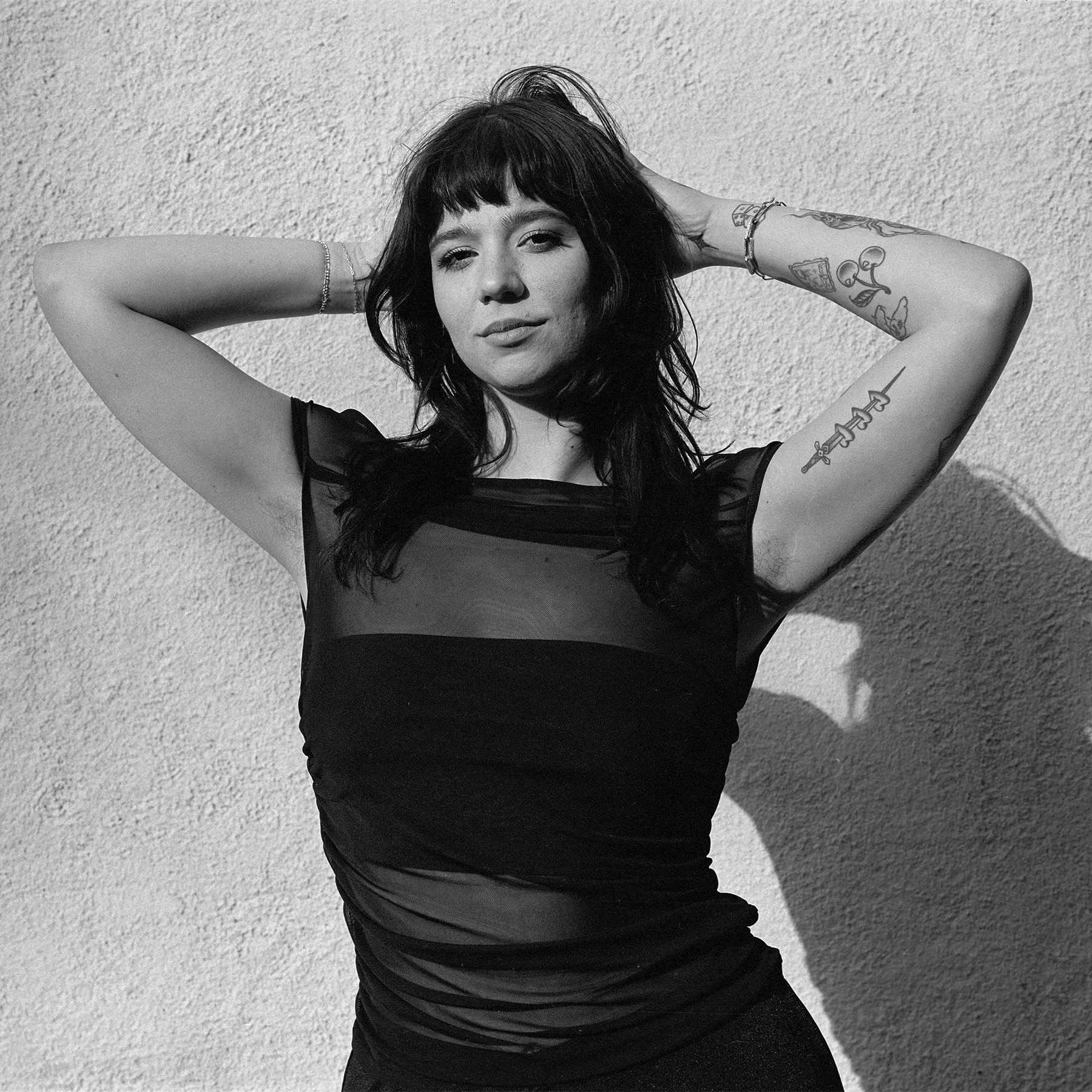 A young person with dark hair and bangs, posing against a textured wall, wearing a black top and showing tattoos on their arms.