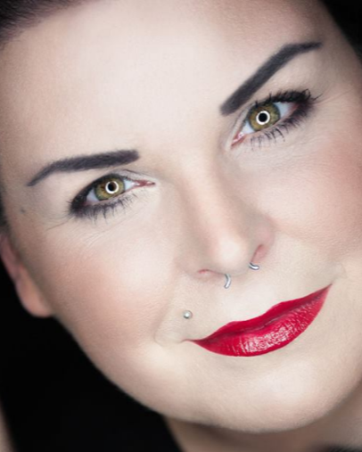 Close-up of a persons face with dark eyebrows, bold eye makeup, a nose ring, a facial piercing, and bright red lipstick.