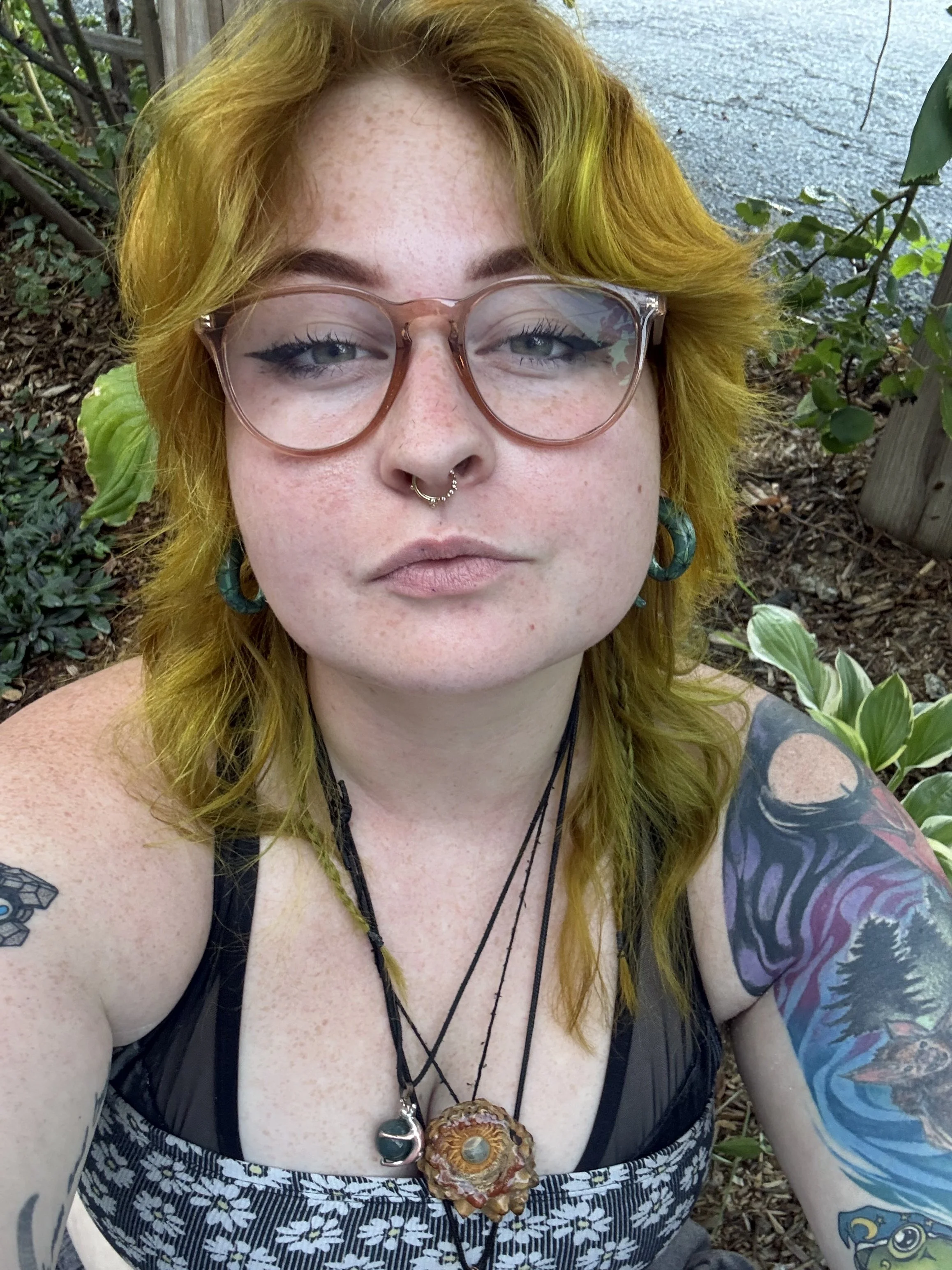 Close-up of a person with yellow hair, wearing glasses, black eyeliner, a nose ring, large turquoise earrings, necklaces, and a patterned dress, outdoors among plants and leaves.