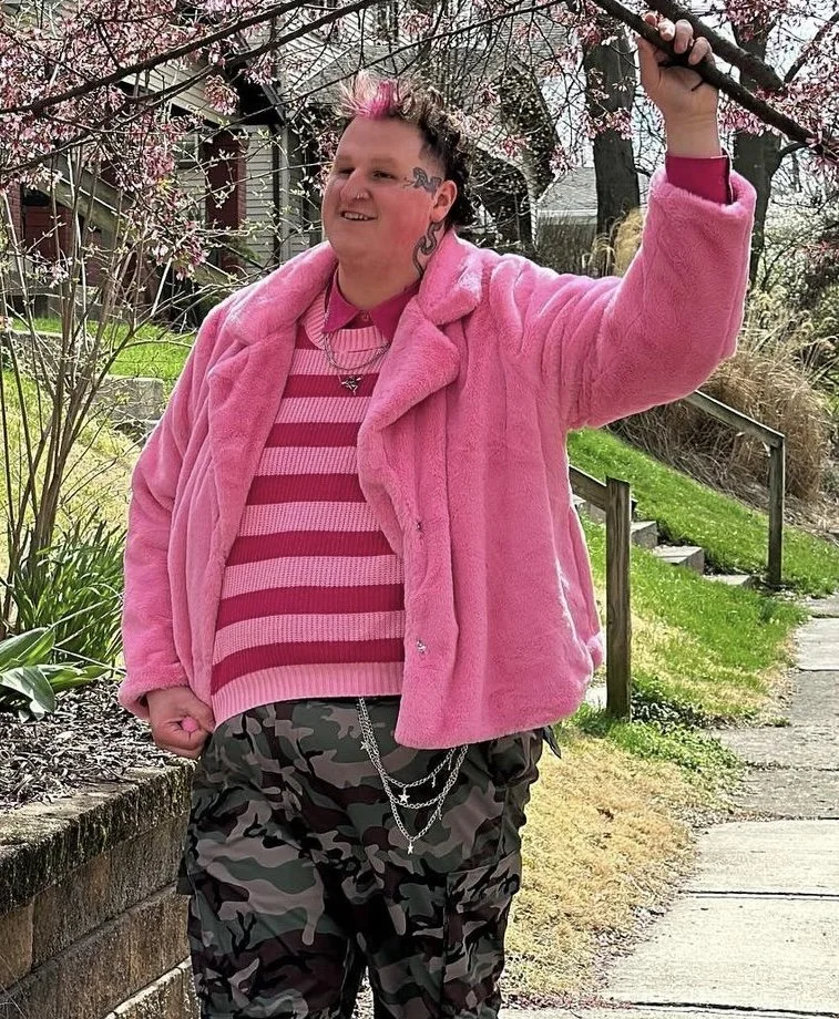 Person with mohawk hairstyle and tattoos on face and neck, wearing a pink fuzzy coat and matching pink striped sweater, standing outdoors on a sidewalk, holding a tree branch with pink blossoms.