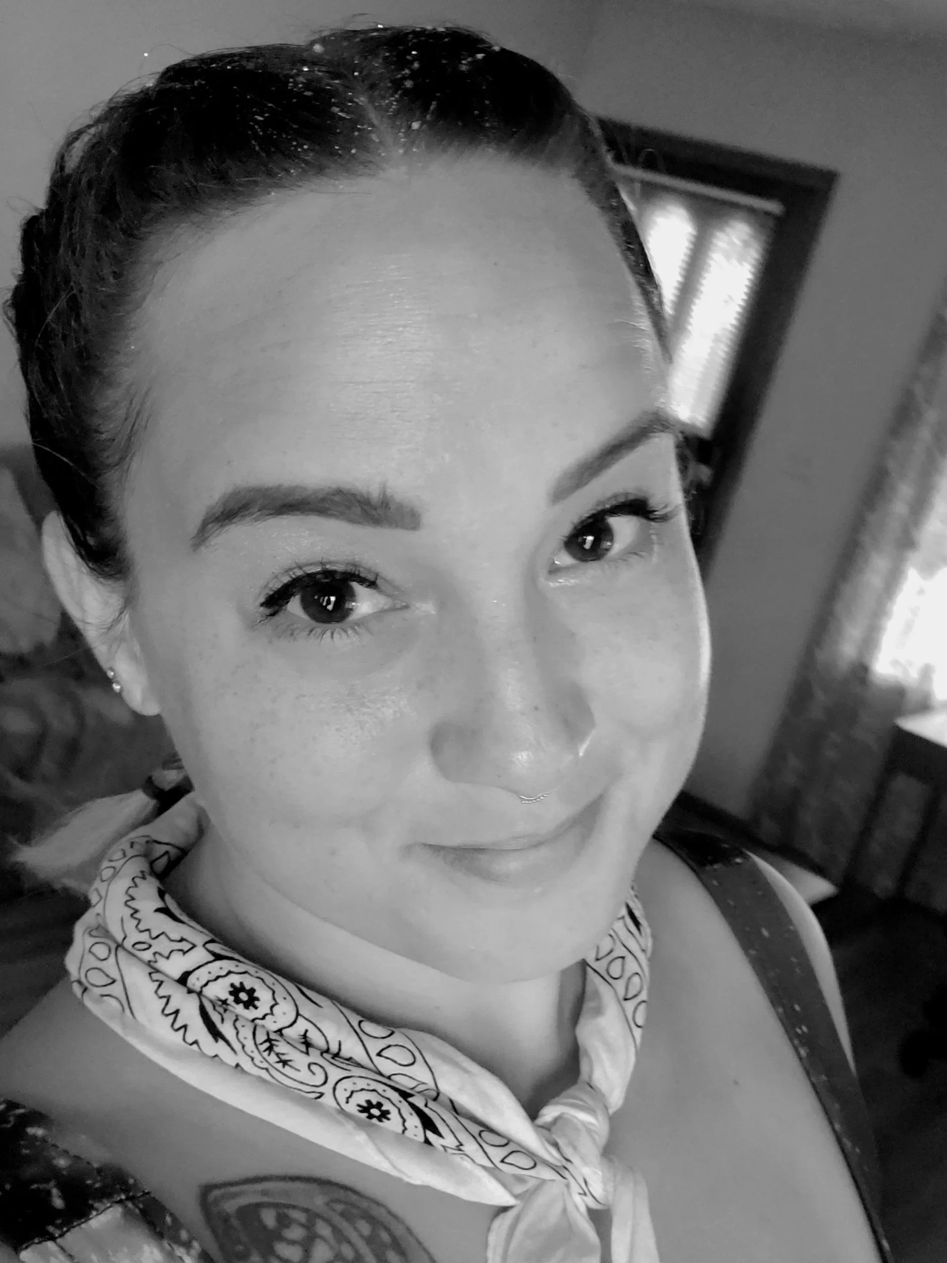 Close-up black and white photo of a person with dark, curled hair tied back, wearing a patterned shirt with a bandana around their neck.