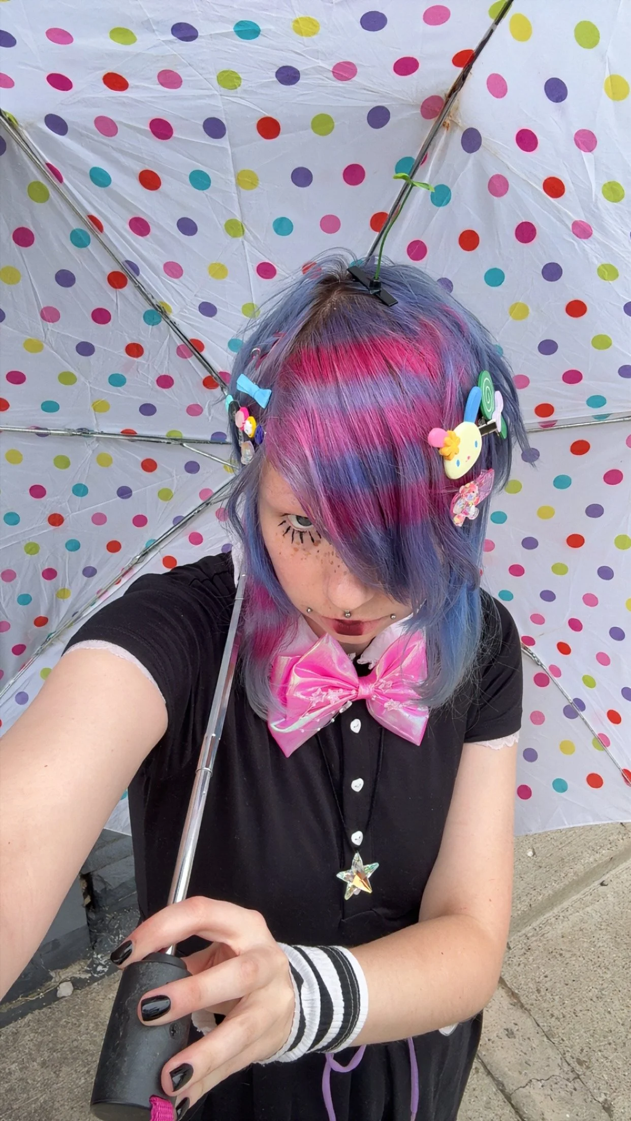 Person with colorful, pastel rainbow hair, decorated with hair clips and makeup, holding a black umbrella with a polka dot pattern, taking a selfie.