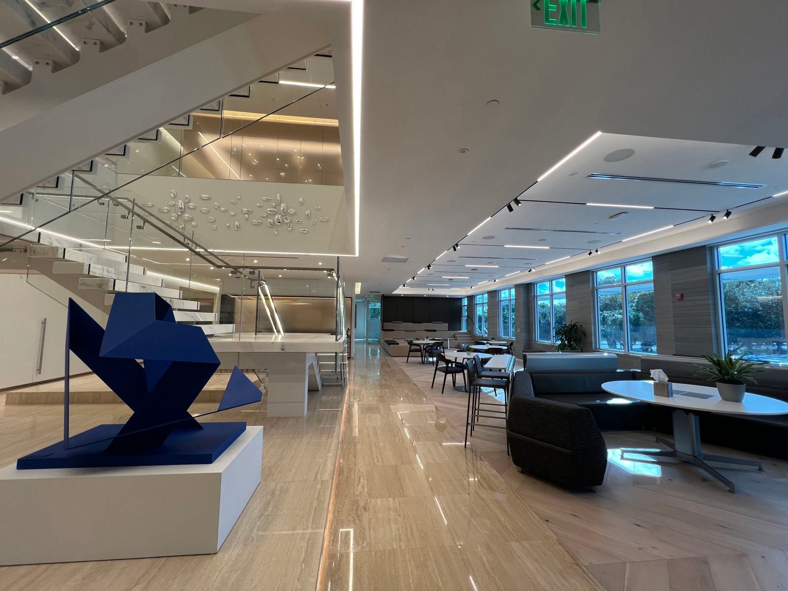 Interior of a modern lounge or lobby area with seating, a staircase, a large blue abstract sculpture on a white pedestal, large windows, and contemporary decor.