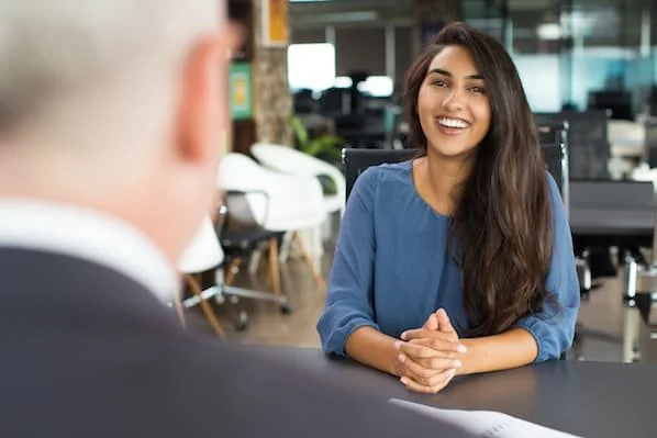 A young woman with long brown hair smiling during a job interview in a modern office environment.