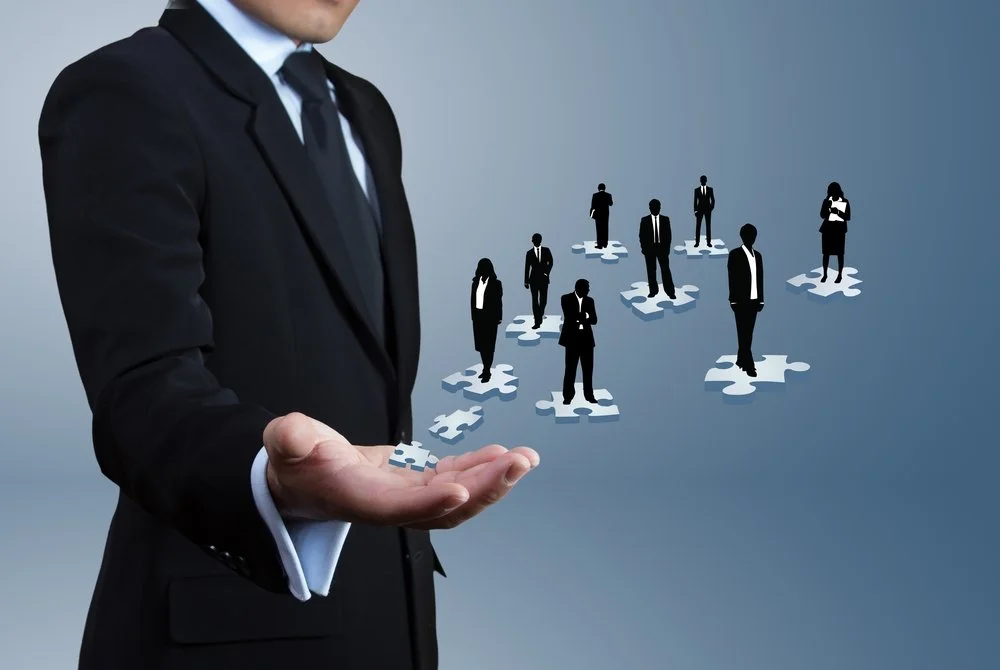 Businessman in suit holding puzzle pieces with silhouettes of businesspeople on puzzle pieces, representing leadership, teamwork and collaboration.