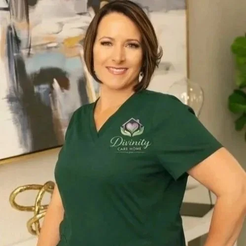 A woman in a green medical scrub top with the logo 'Divinity Care Home' on it, smiling in a modern, decorated room.