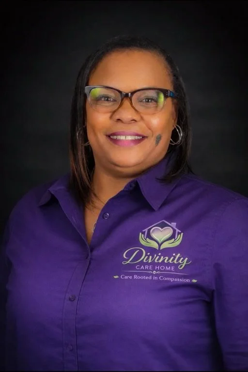 A woman with glasses and dark hair smiling, wearing a purple shirt with the logo of Divinity Care Home.