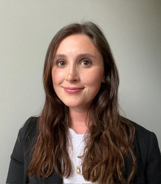 Portrait of a woman with long wavy brown hair, smiling, wearing a black blazer over a white top, standing against a plain light-colored wall.