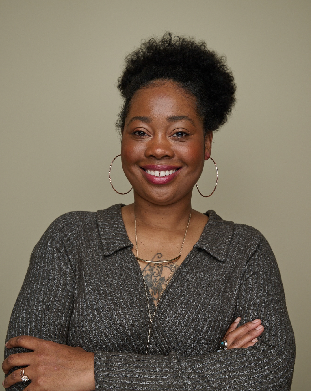 A smiling African American woman with curly hair, wearing hoop earrings and a dark gray sweater, standing against a beige background.
