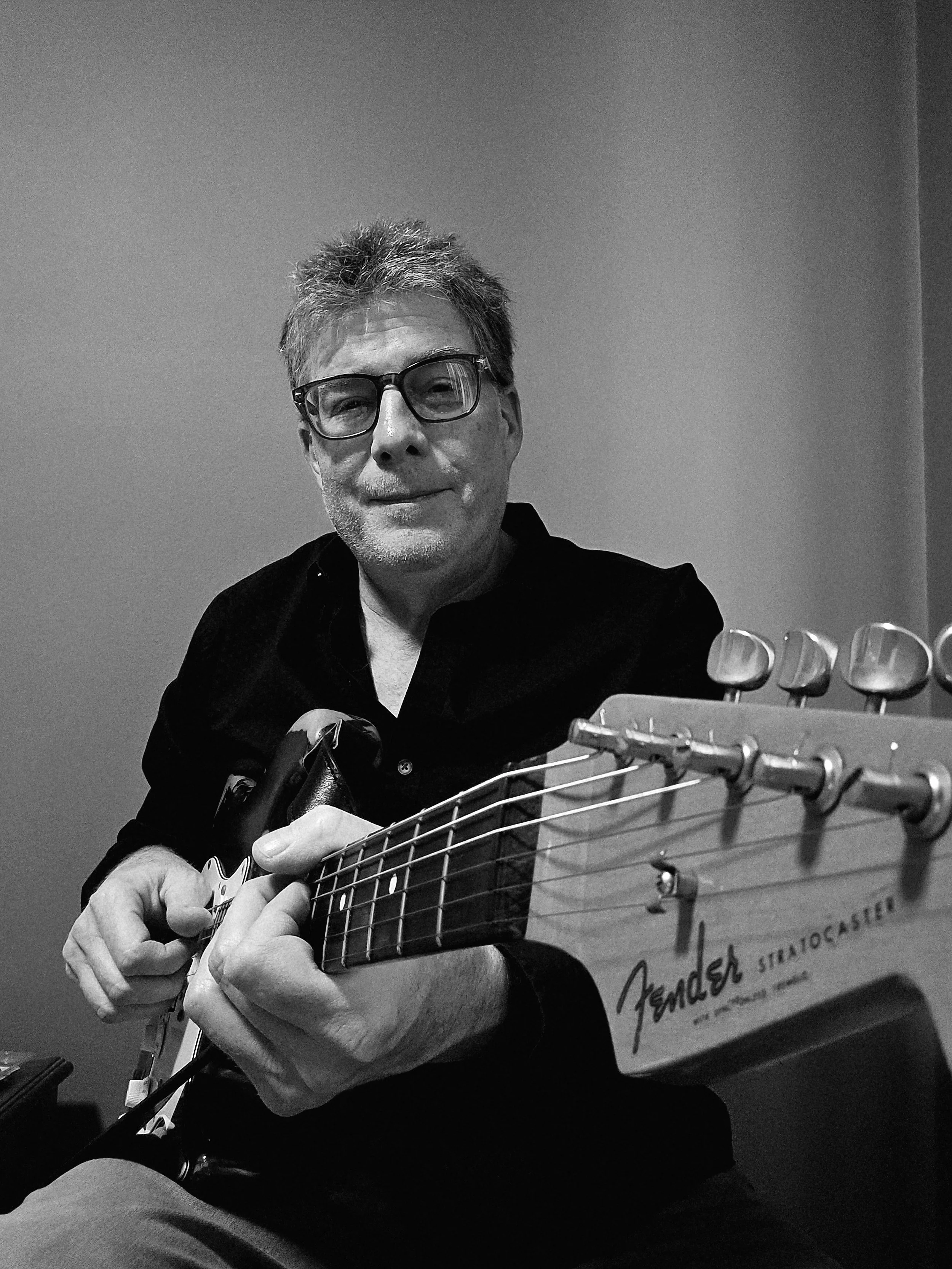 A man with glasses playing an electric guitar, with a plain wall background.