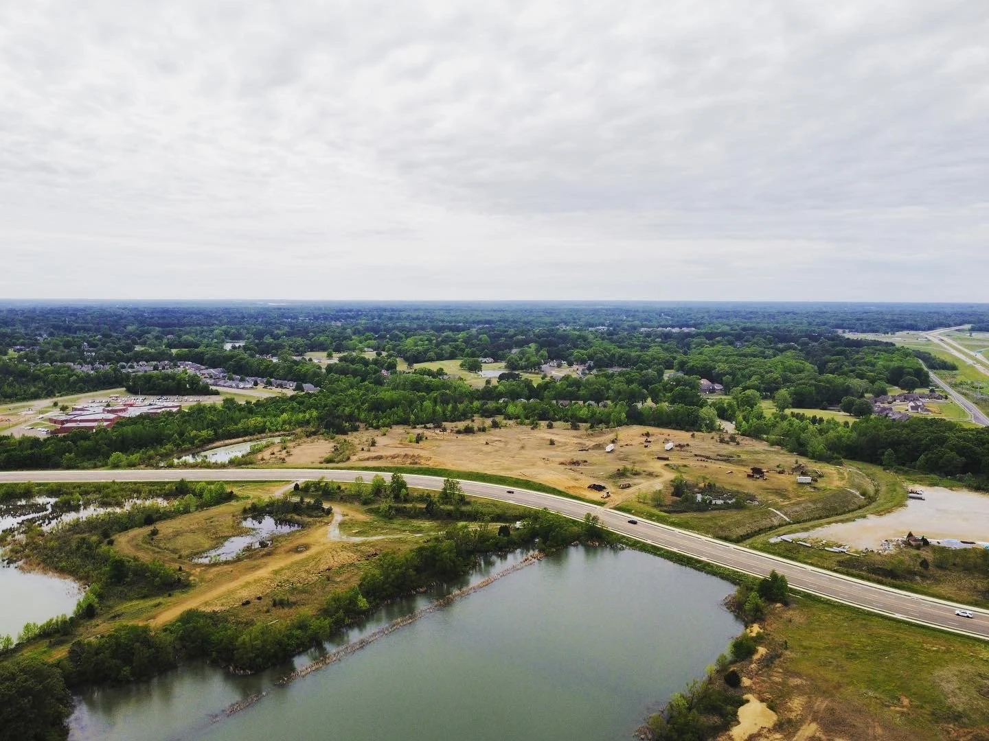 Aerial view of a rural landscape with water bodies, a highway, grassy fields, trees, and scattered houses. Overcast sky.
