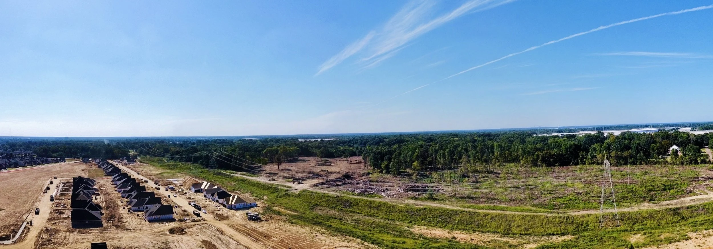 Aerial view of a developing neighborhood with houses under construction, surrounded by greenery and trees, under a blue sky with scattered clouds.