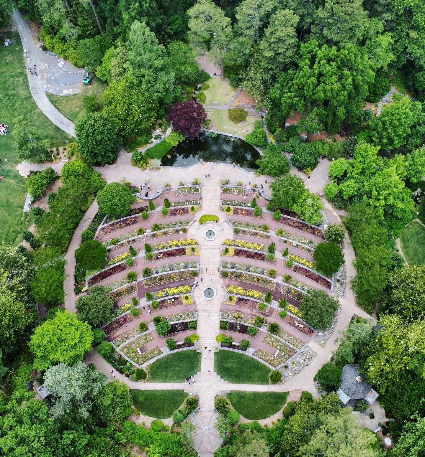 An aerial view of a landscaped park with pathways, flower beds, a pond, and lush green trees.
