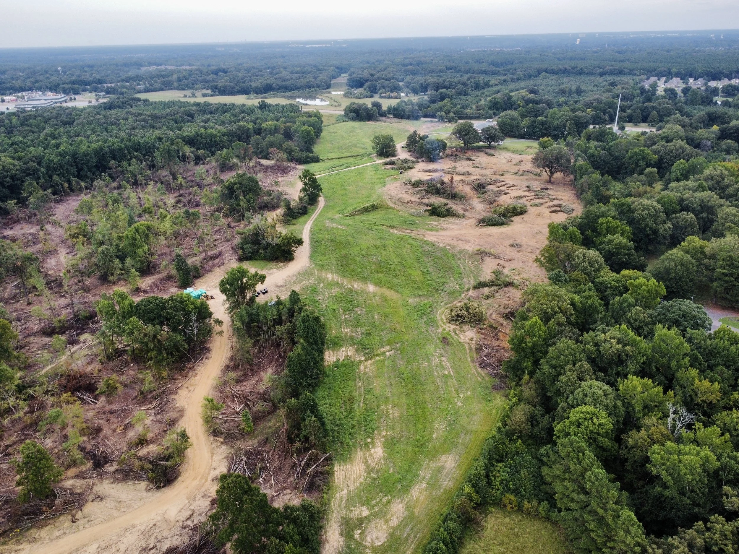 Aerial view of land with cleared areas, green grassy patches, and surrounding dense trees, with a dirt road winding through the landscape.
